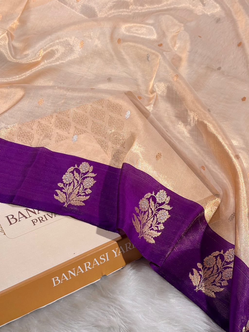 A close-up of a draped Banarasi silk saree in beige with a rich purple border featuring gold floral motifs, resting on a white faux fur surface next to a book.