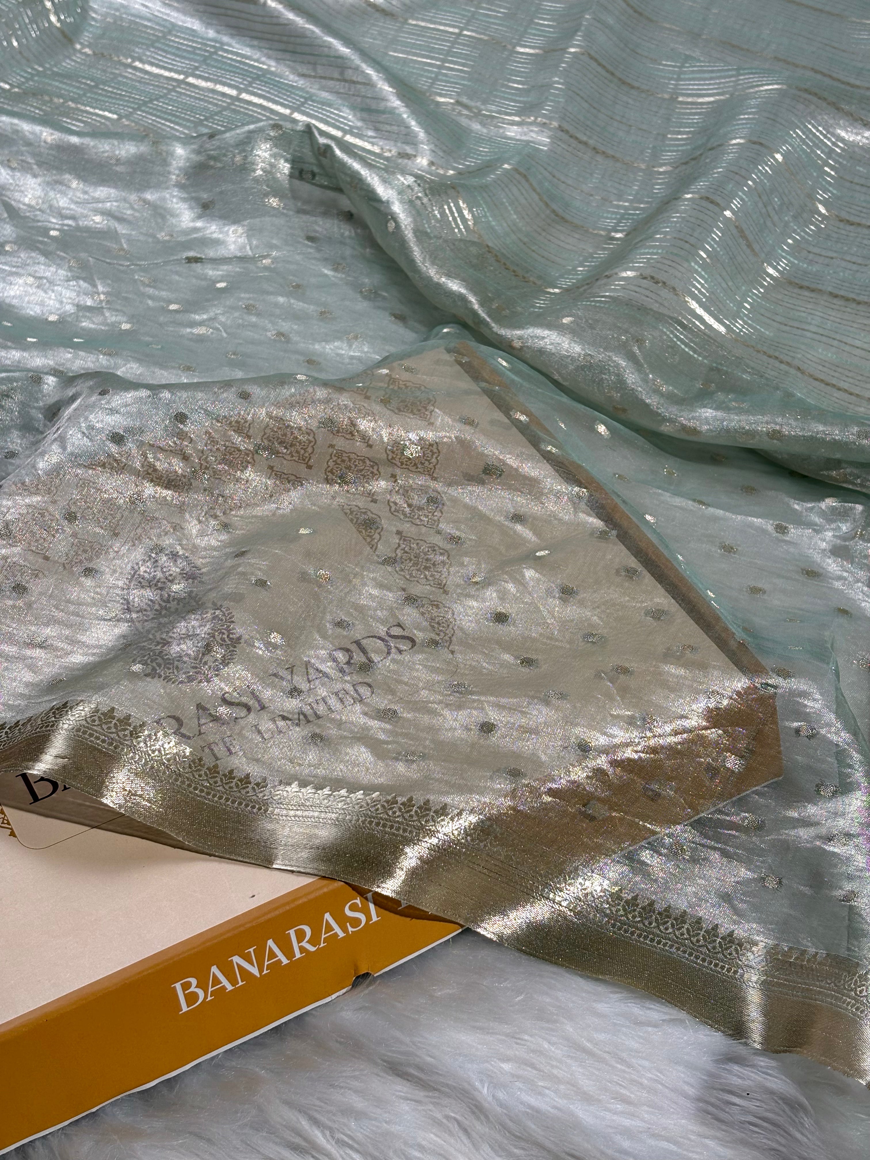 A close-up of a sheer mint green saree with subtle gold zari stripes, a white faux fur surface. The Banarasi Yards box is partially visible underneath.