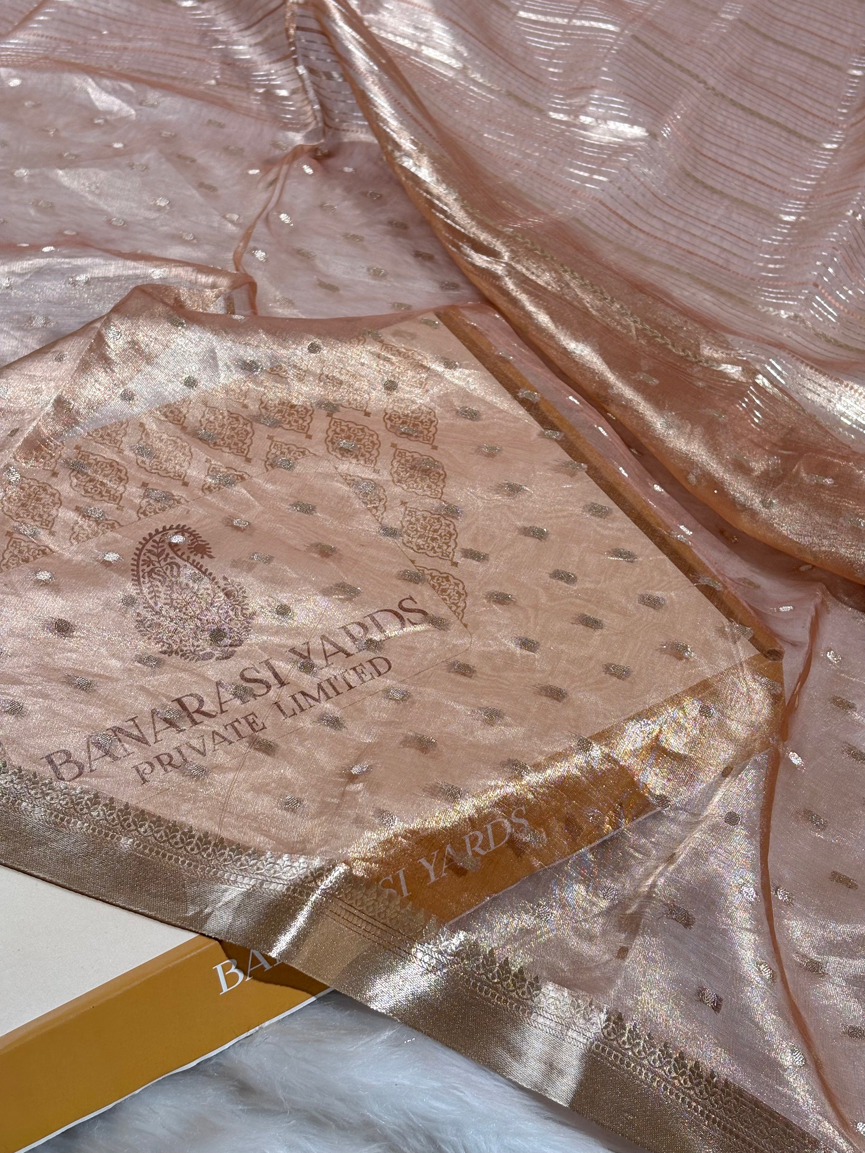 A close-up of a peach-beige sheer organza saree with metallic gold zari and a printed Banarasi Yards Private Limited logo.