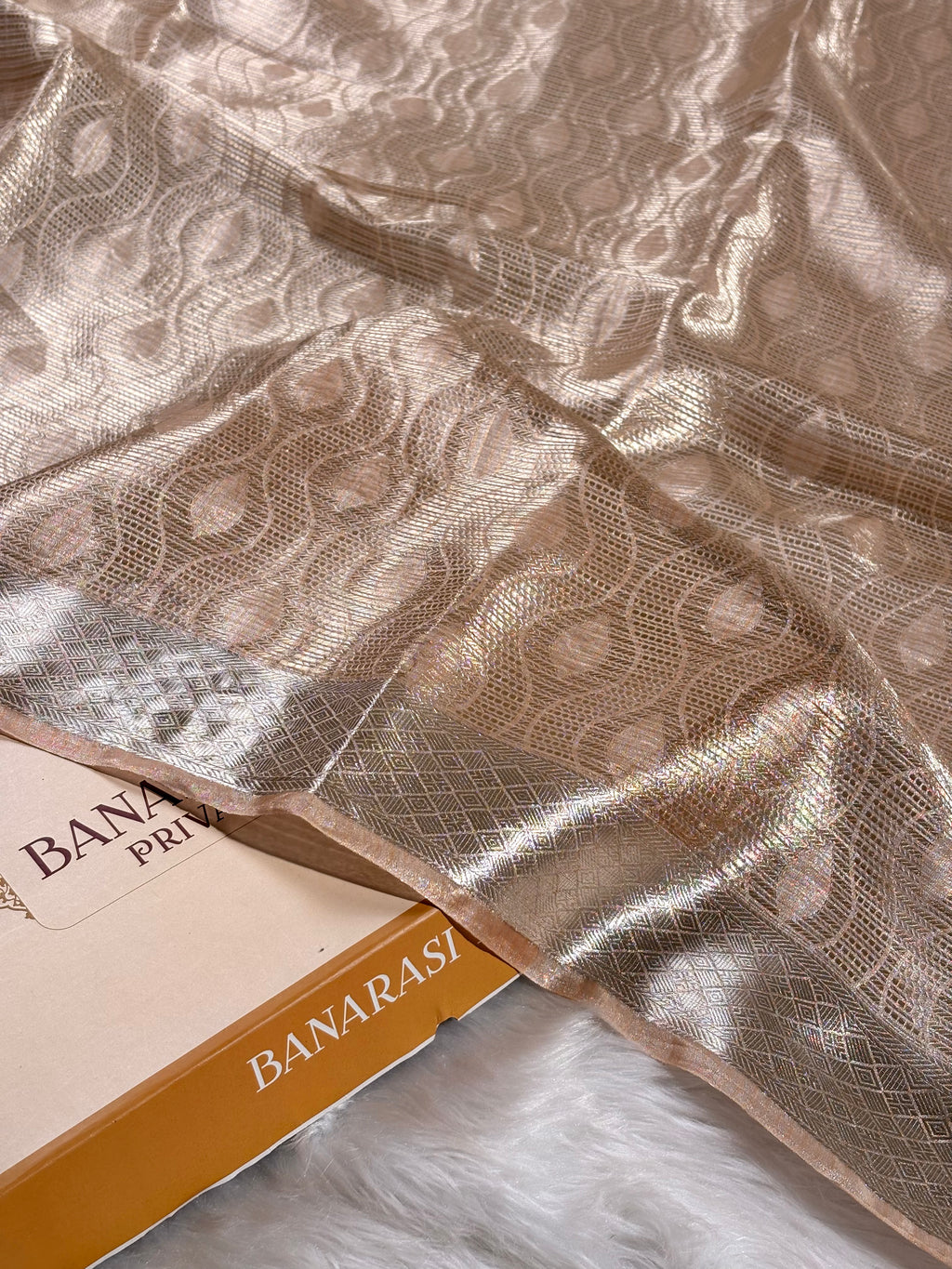 A close-up of a metallic champagne-gold saree with an allover woven circular pattern and a contrasting border, draped over a box labeled 'BANARASI YARDS' on a white faux-fur surface.