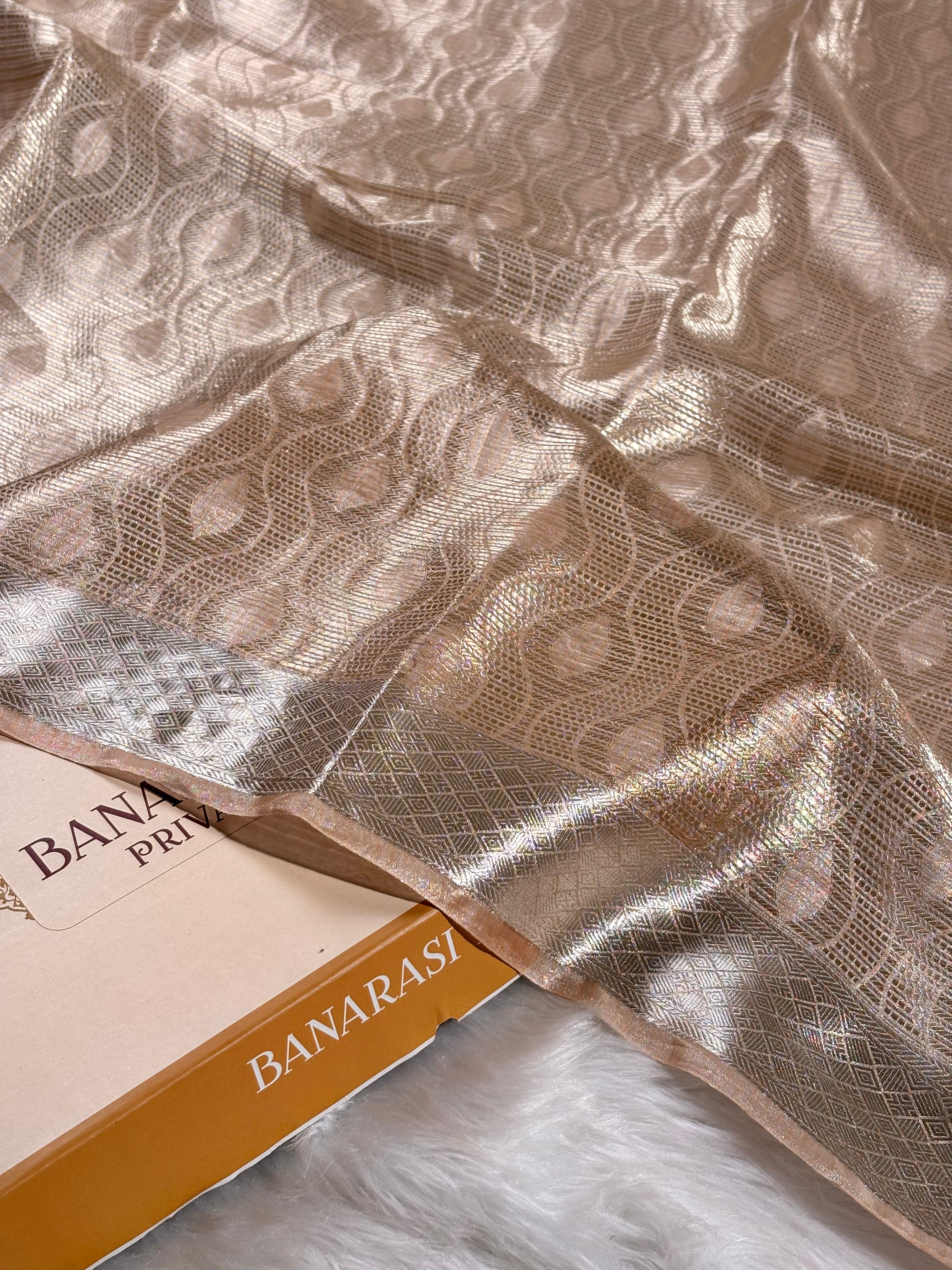 A close-up of a metallic champagne-gold saree with an allover woven circular pattern and a contrasting border, draped over a box labeled 'BANARASI YARDS' on a white faux-fur surface.