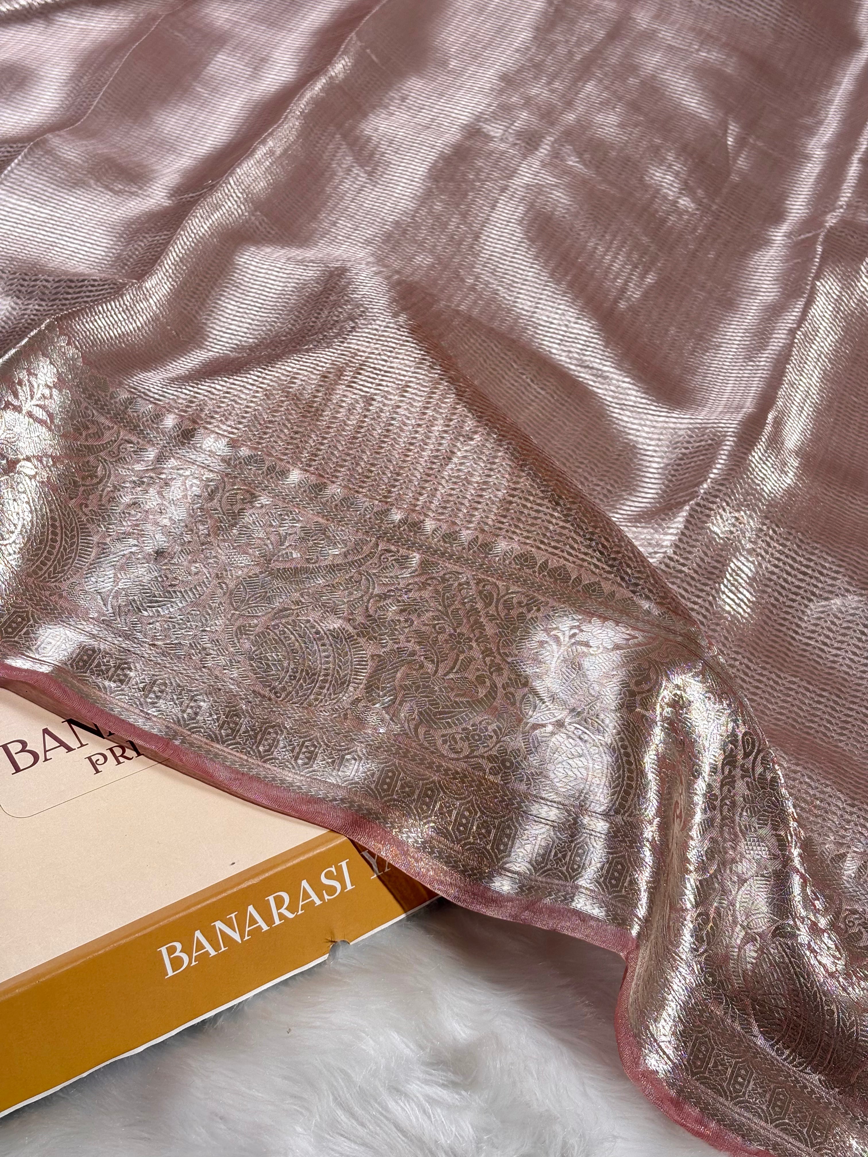 A close-up of a shiny pink Banarasi silk saree with a detailed metallic silver border, draped over a book labeled 'BANARASI YARDS'.