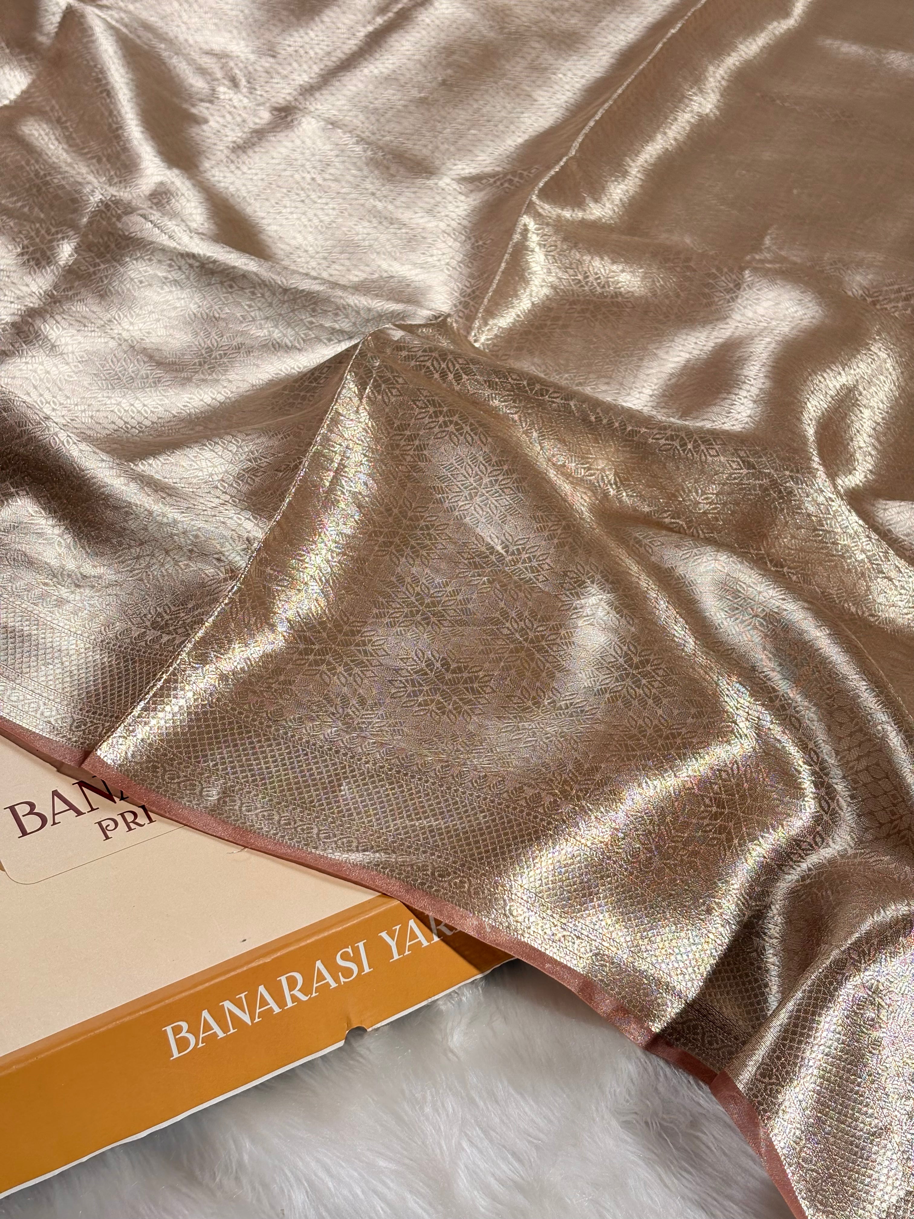 A close-up of draped metallic Banarasi silk saree with a subtle woven pattern, resting on a white faux fur surface next to a box titled 'BANARASI YARDS'.