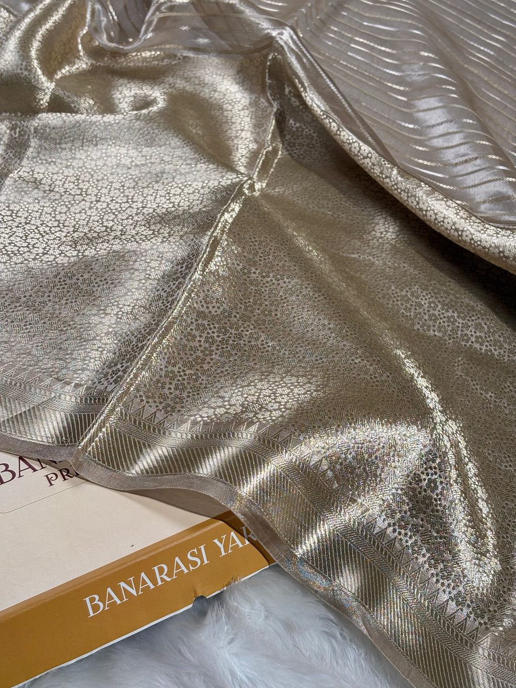 A close-up of shimmering metallic saree draped across a surface, with a folded section showing a fine woven pattern and a gold-toned edge. Part of a book or box labeled BANARASI YARDS is visible underneath.