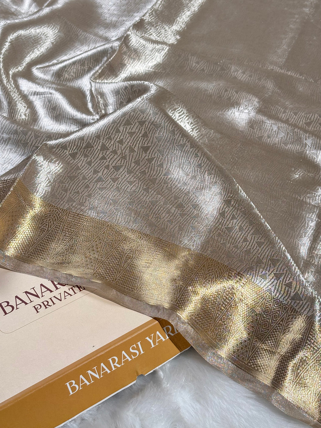 A close-up of a Banarasi silk saree with a metallic silver body, a gold border, and a repeating woven pattern, draped over a white faux fur surface.