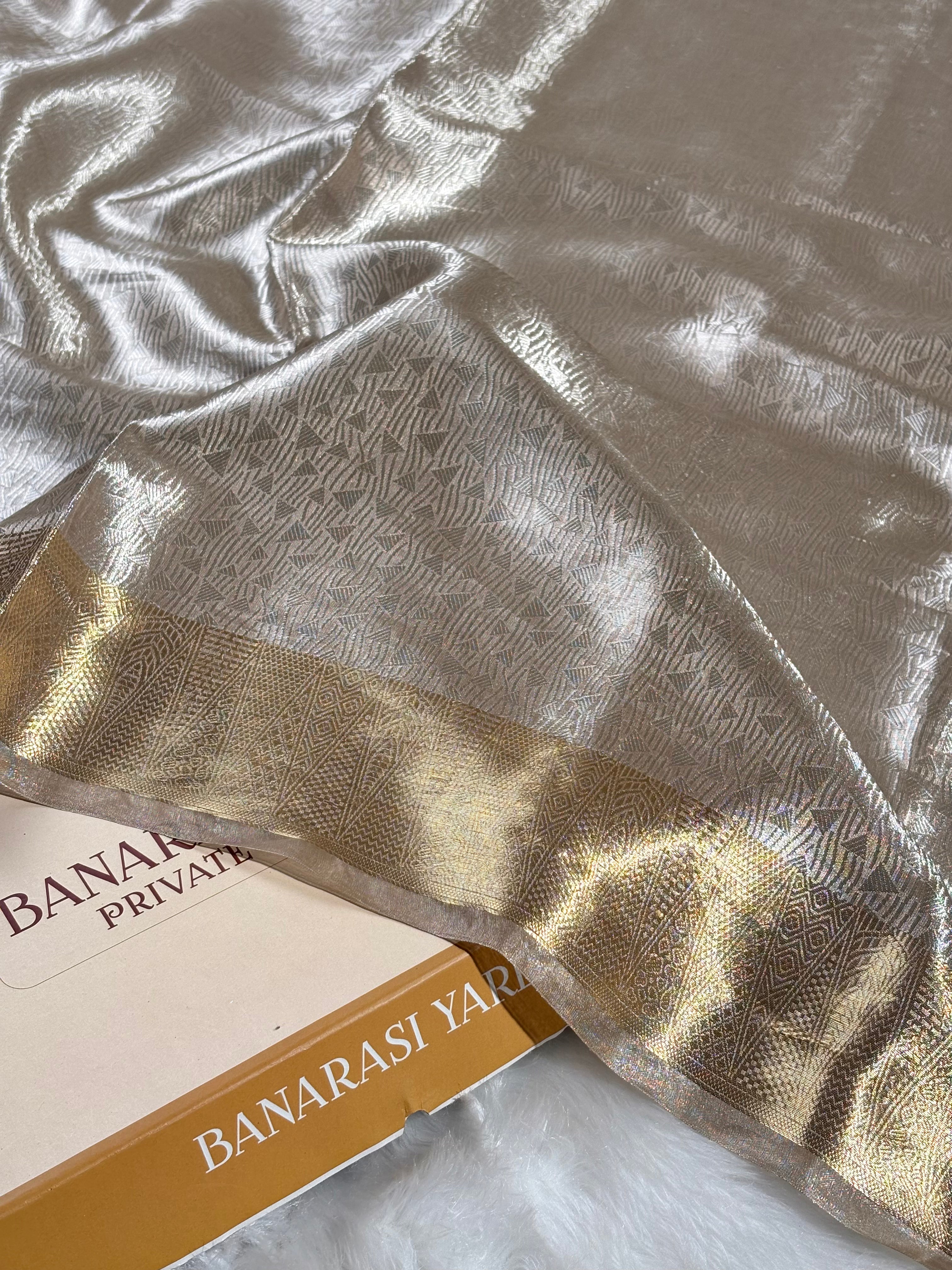 A close-up of a Banarasi silk saree with a metallic silver body, a gold border, and a repeating woven pattern, draped over a white faux fur surface.