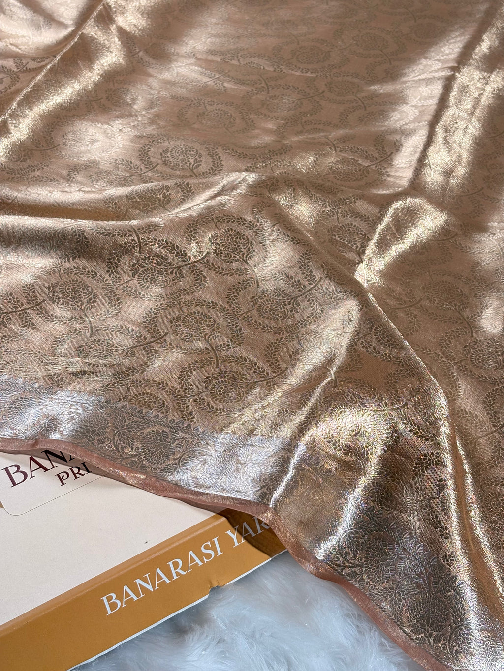A close-up of draped metallic gold Banarasi silk saree with an intricate woven floral pattern, resting on a white faux fur surface next to a box labeled 'BANARASI YARDS.