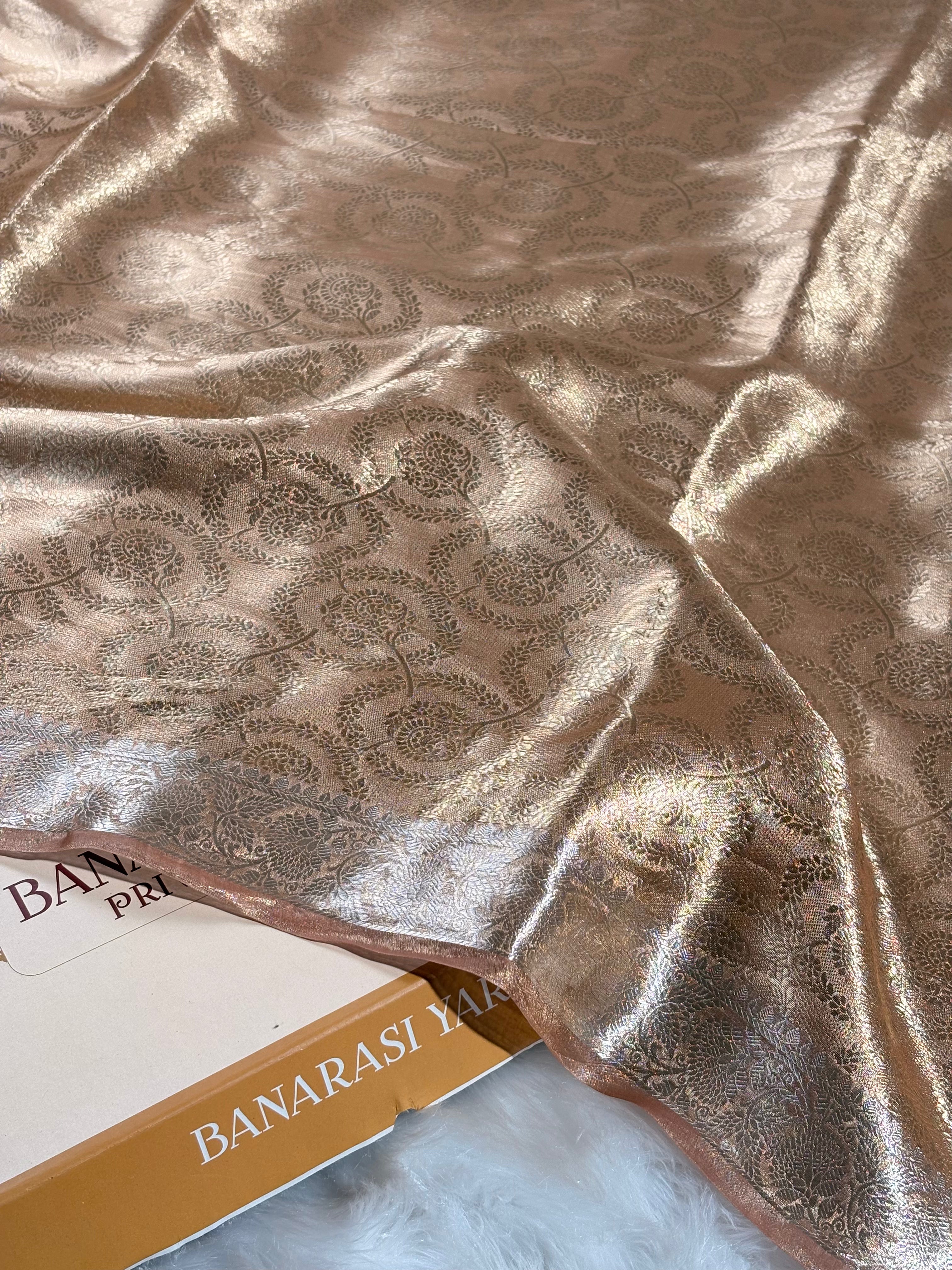 A close-up of draped metallic gold Banarasi silk saree with an intricate woven floral pattern, resting on a white faux fur surface next to a box labeled 'BANARASI YARDS.