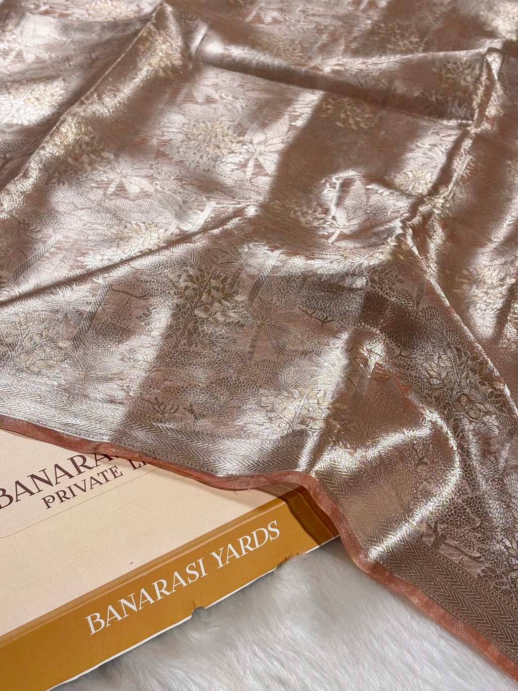 A close-up of a metallic rose gold Banarasi silk saree with subtle floral zari weaving, draped elegantly over a Banarasi Yards box on a white faux fur surface.