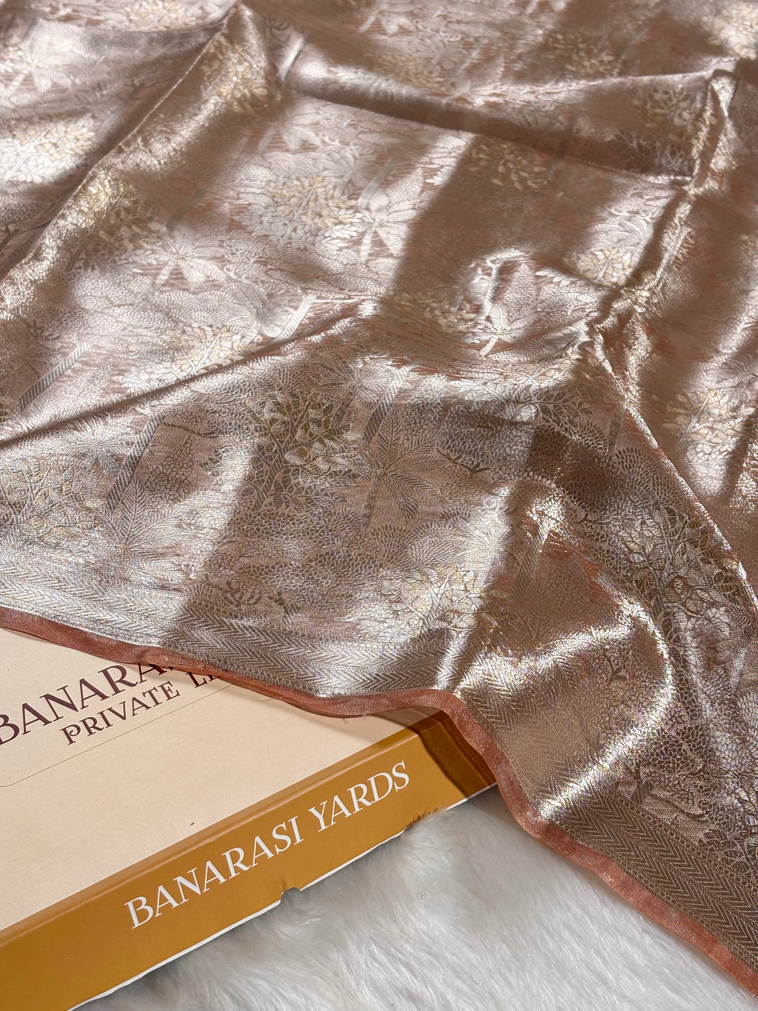A close-up of a metallic rose gold Banarasi silk saree with subtle floral zari weaving, draped elegantly over a Banarasi Yards box on a white faux fur surface.