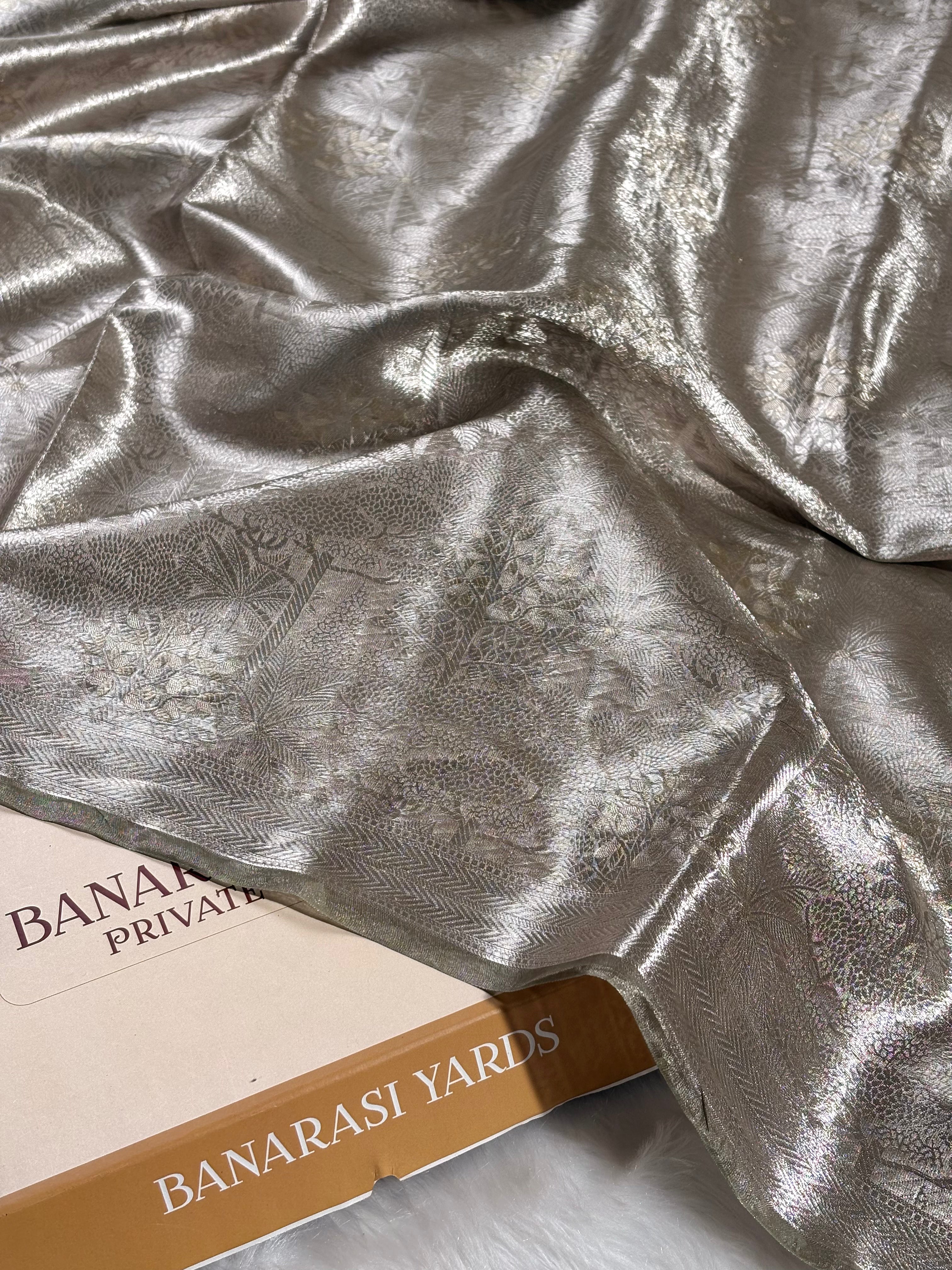 A close-up of draped metallic silver Banarasi saree with a subtle floral brocade pattern, resting on top of a beige and brown paper box labeled 'BANARASI YARDS'.