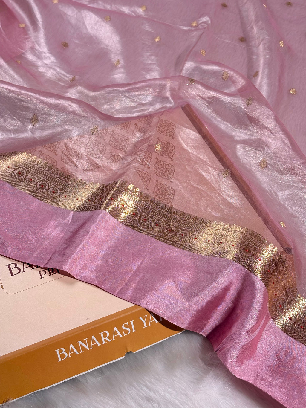 A close-up of a pink Banarasi silk saree with a wide metallic gold border and scattered small gold motifs, draped over a box labeled 'BANARASI YARDS'.