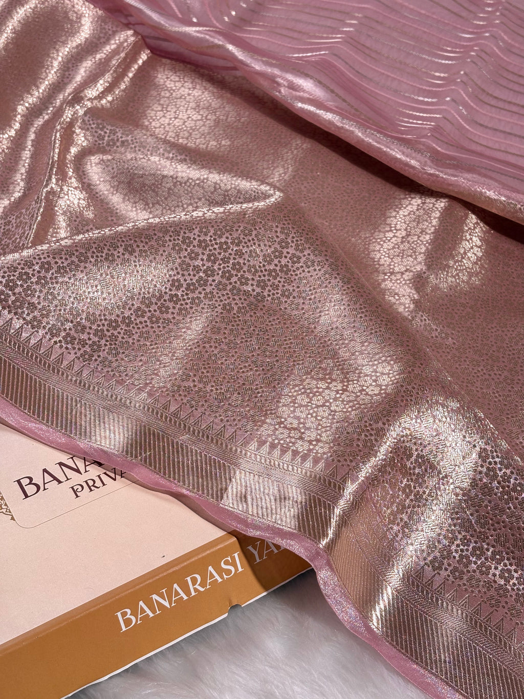 A close-up of a metallic pink Banarasi silk saree draped elegantly, showing its shimmering texture and border, with a Banarasi Yards Private Limited label box visible underneath.