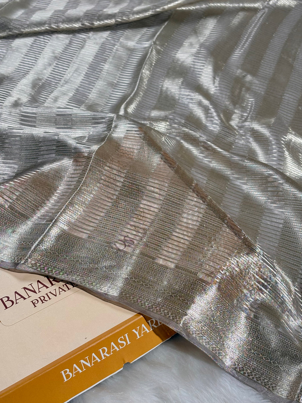 A close-up of a metallic silver Banarasi silk saree with a striped pattern and a decorative border, draped over a book labeled 'BANARASI YARDS'.