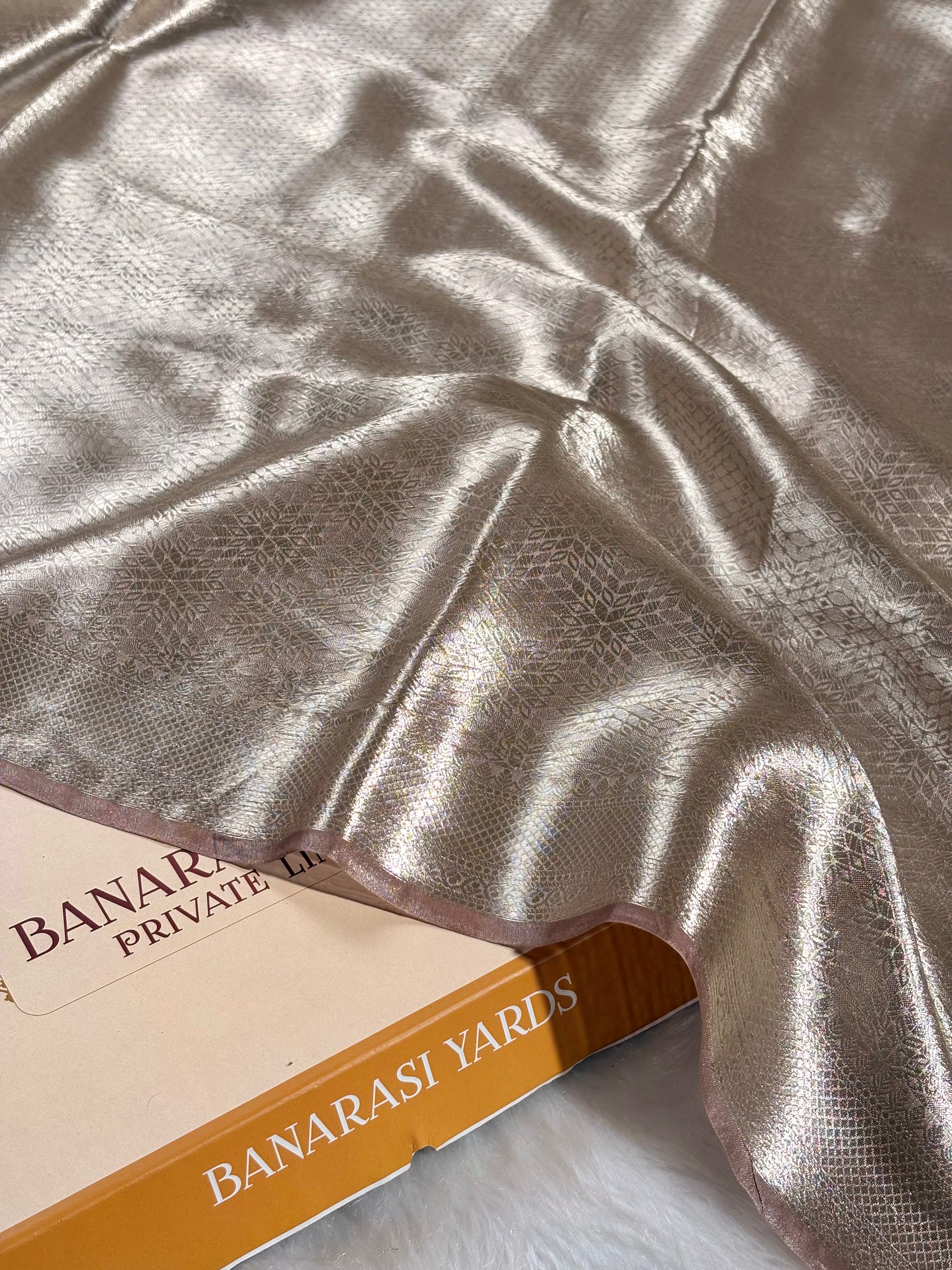 A close-up of pure molten tishu banarasi saree draped over a box labeled BANARASI YARDS. The saree has a reflective silver-gold sheen with a subtle woven texture.