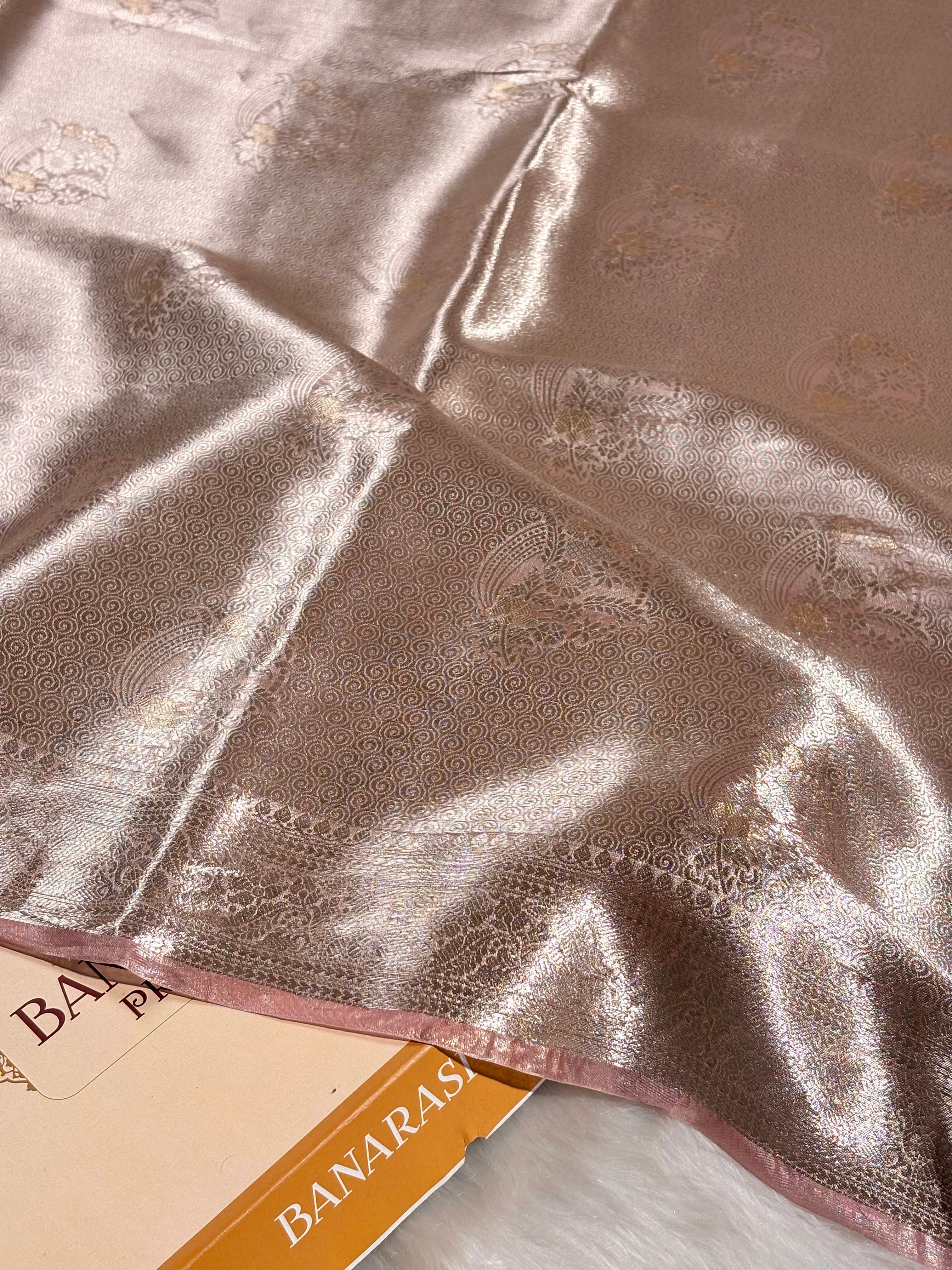 A close-up of a pure molten tishu saree in a warm taupe-beige color with subtle woven floral motifs and a decorative border, draped over a book labeled BANARASI YARDS.