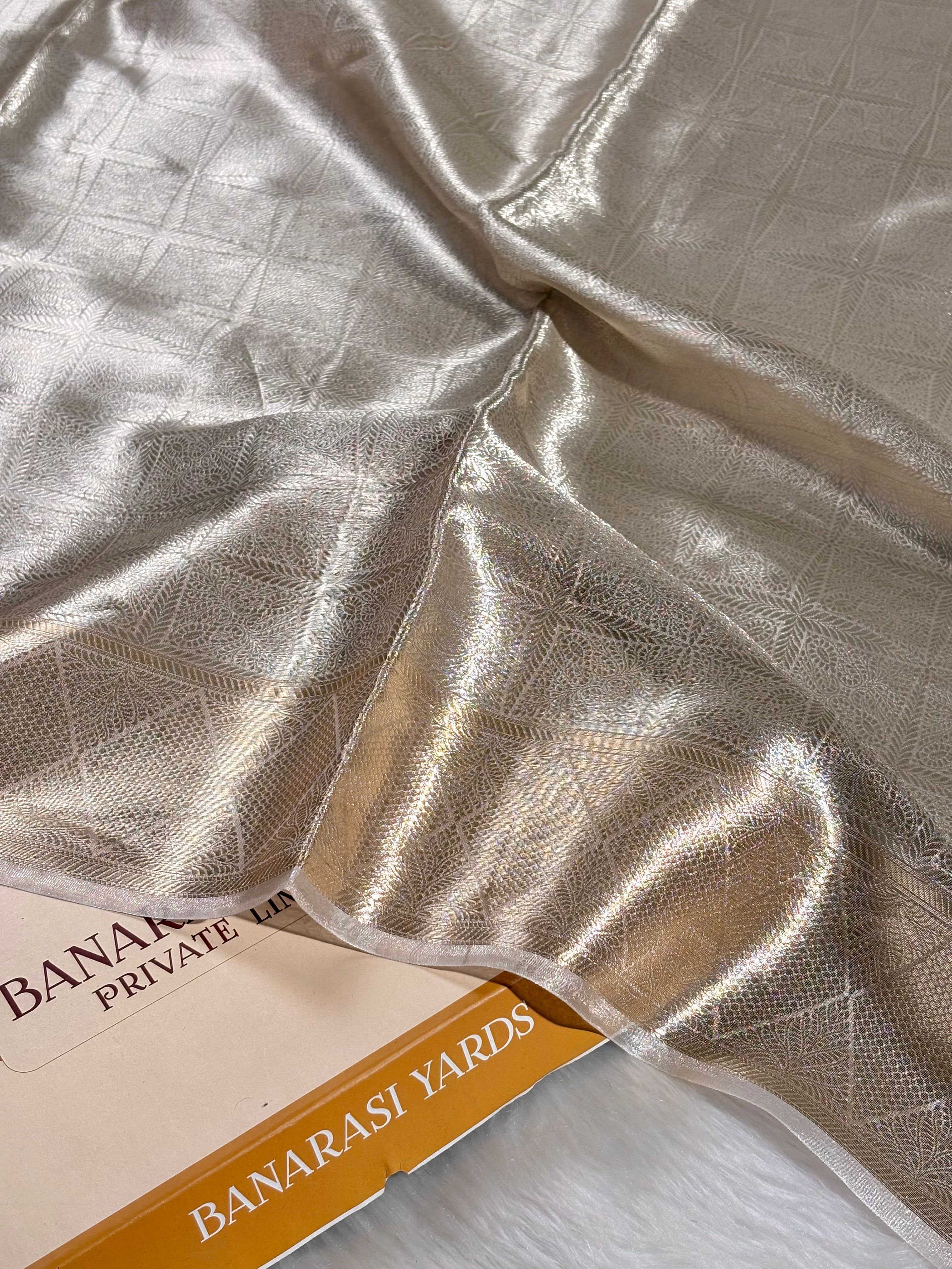 A close-up of draped metallic silver saree with a subtle woven pattern, partially covering a book or box labeled 'BANARASI YARDS'.