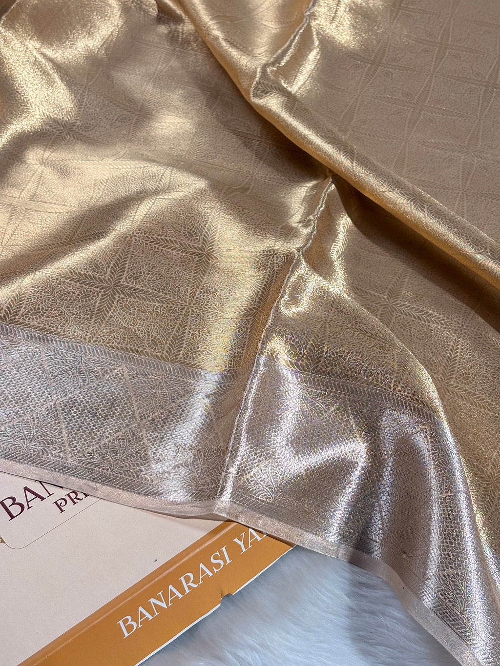 A close-up of draped metallic gold Banarasi silk saree with a subtle woven geometric pattern and a shimmering border, resting on a white faux fur surface next to a box.