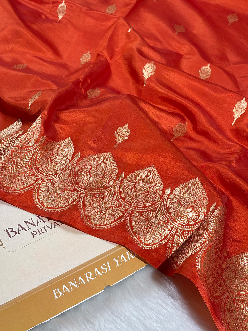 A close-up of a rich red-orange silk saree with intricate gold zari work, featuring a decorative border and scattered metallic motifs, draped over a white surface with a branded box partially visible.