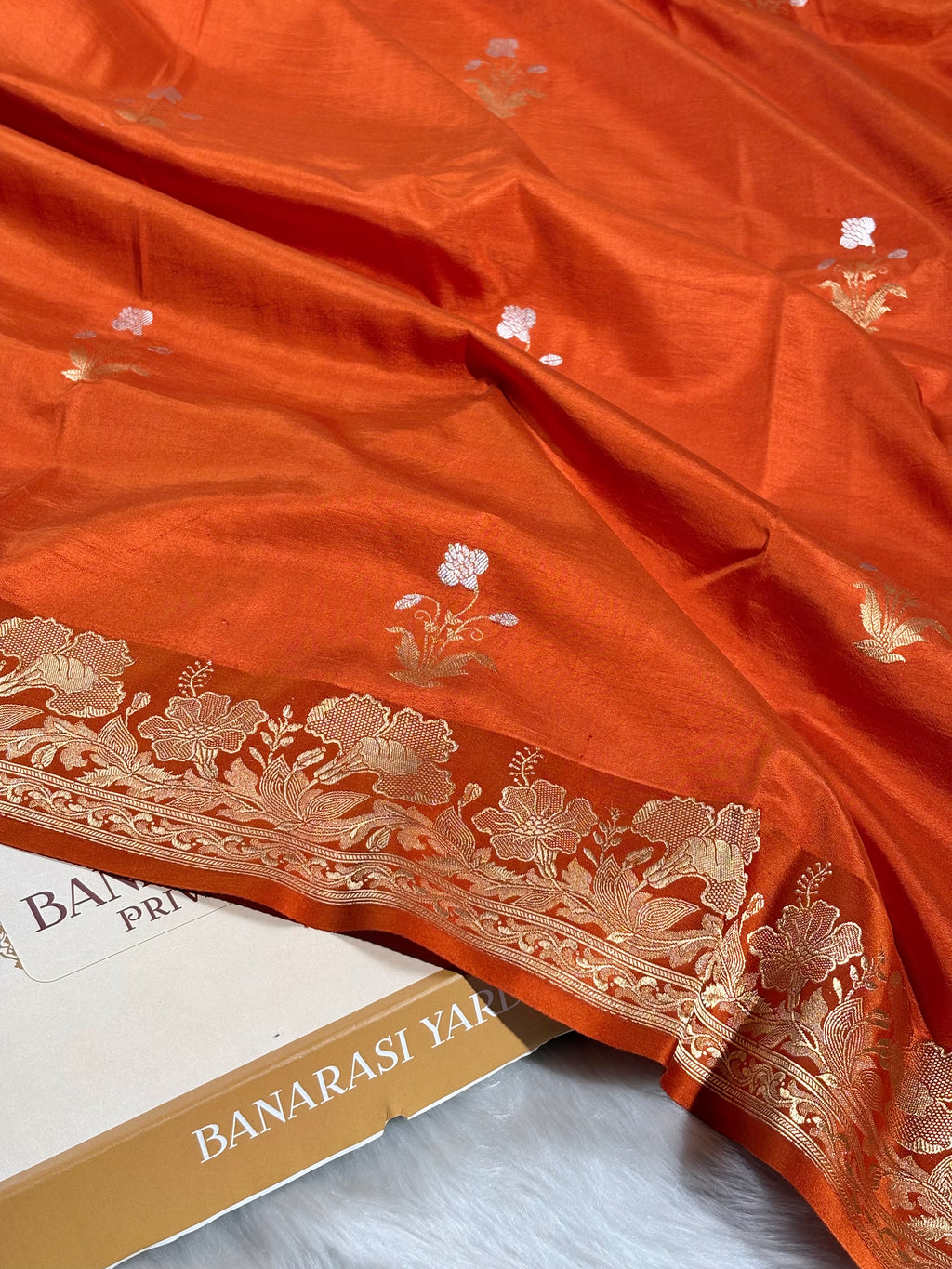 An orange Banarasi silk saree with a wide gold floral border, draped elegantly over a box labeled 'BANARASI YARDS'.