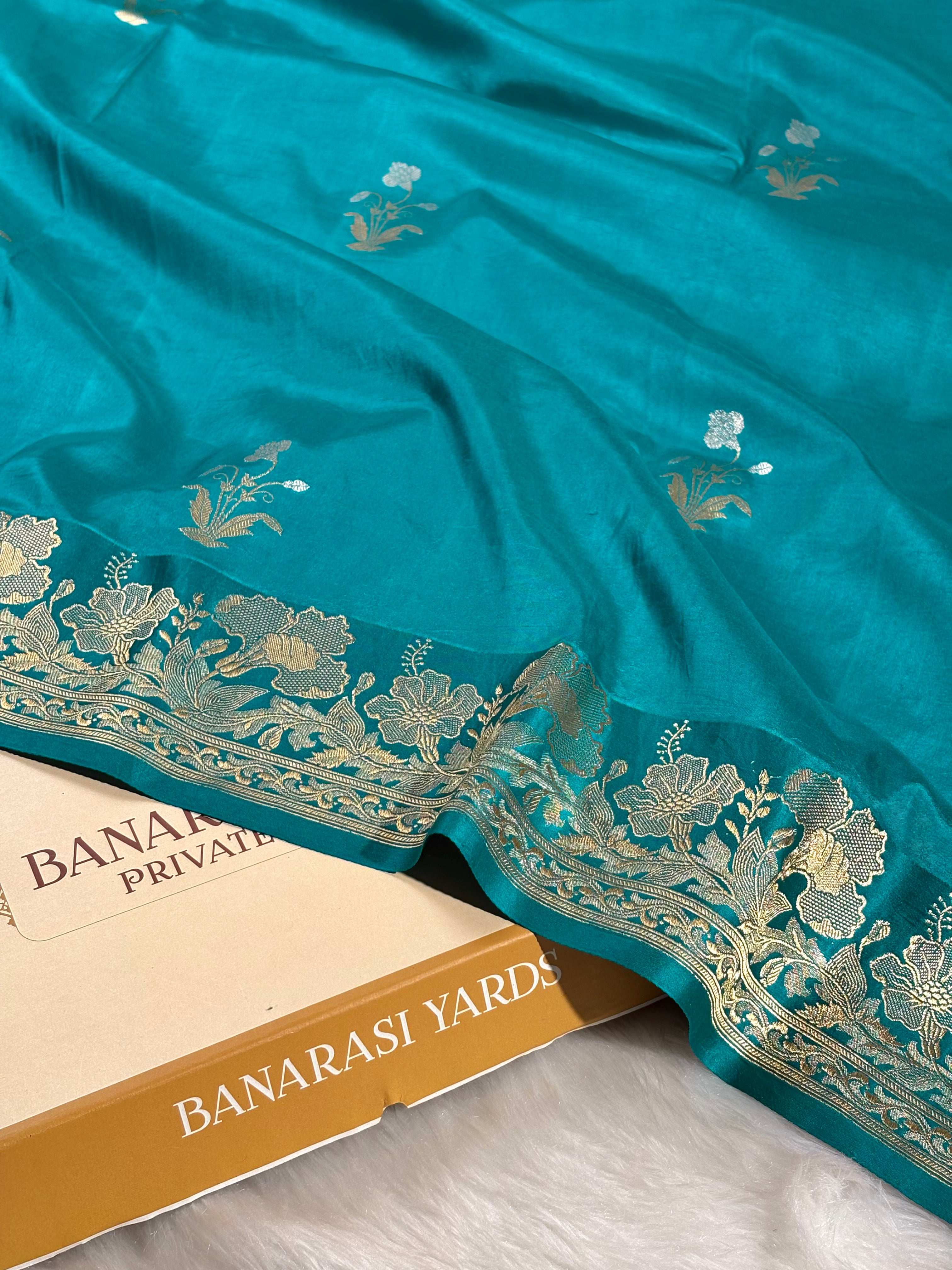 A teal blue Banarasi silk saree with intricate alfi mina floral weaving along the border, draped elegantly over a Banarasi Yards label box on a white faux fur surface.