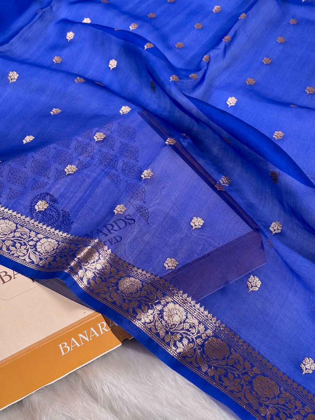 A royal blue silk saree with intricate gold zari woven borders and small scattered gold motifs, draped elegantly over a matching blue saree, with part of a box visible at the bottom left.