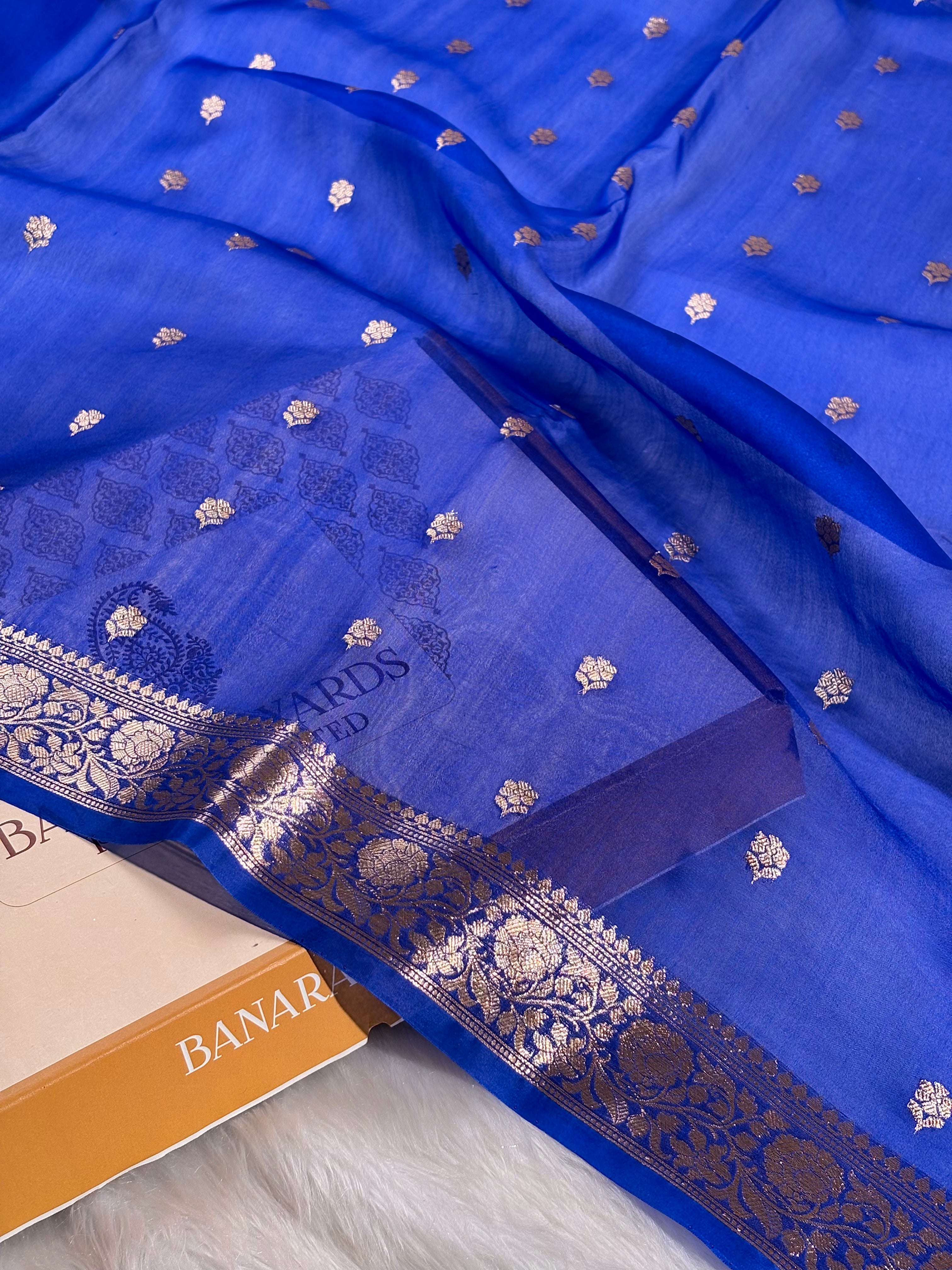 A royal blue silk saree with intricate gold zari woven borders and small scattered gold motifs, draped elegantly over a matching blue saree, with part of a box visible at the bottom left.