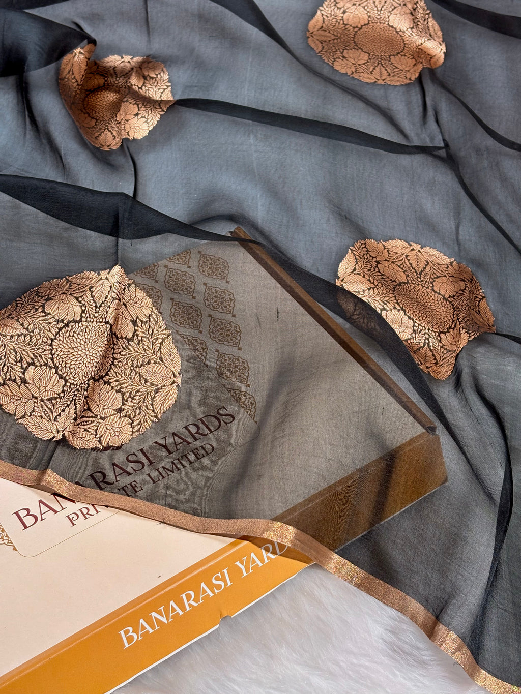 A draped slate blue-grey saree with beige floral weaving, shown with its branded Banarasi Yards box and paper packaging on a white faux-fur surface.