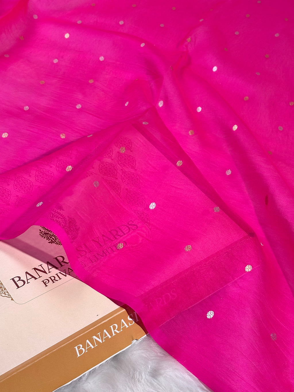Bright pink Banaras silk saree with small metallic alfi mina dot patterns, draped elegantly with a matching paper box and a white faux fur surface.