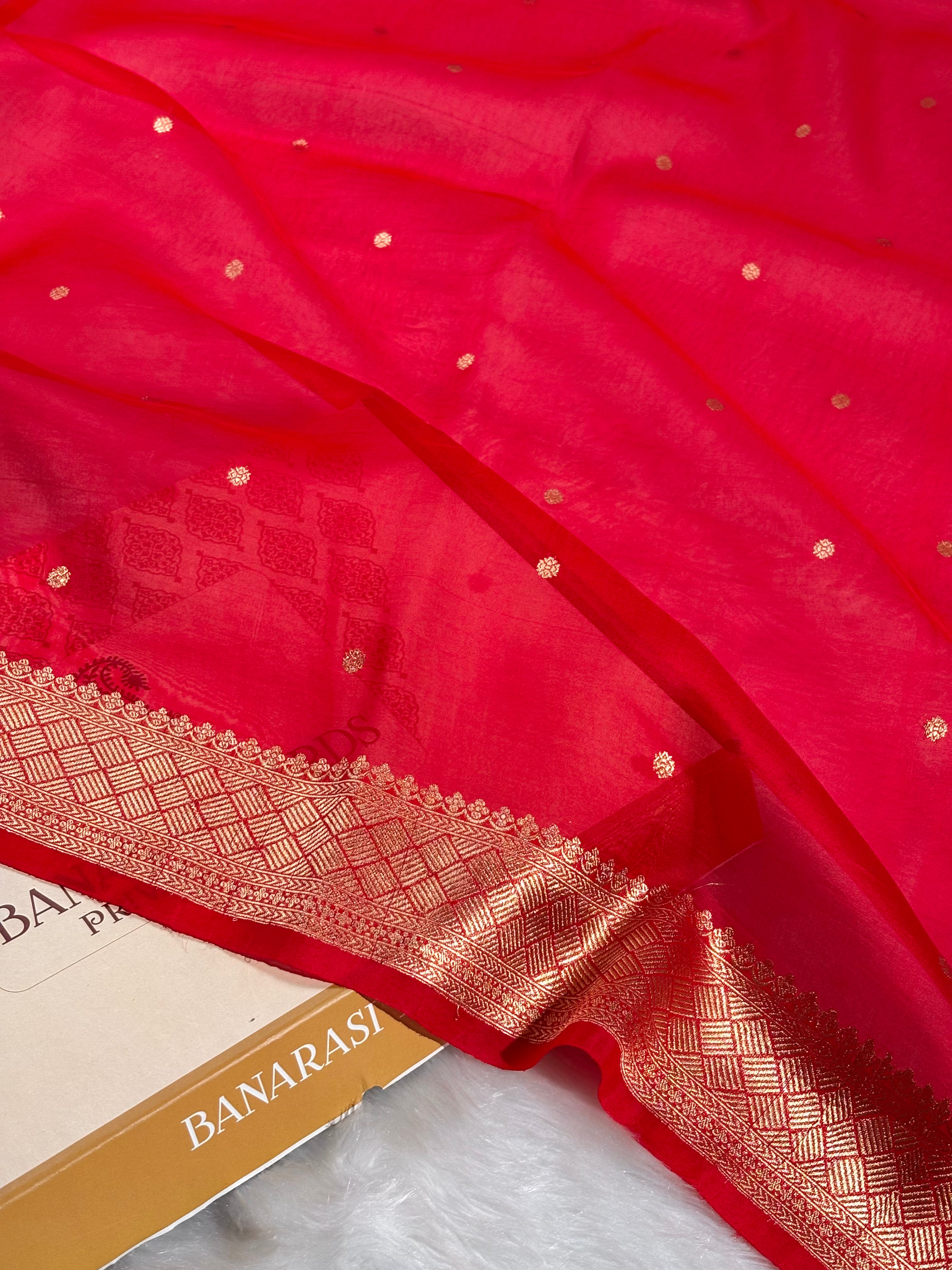 A close-up of a bright red sheer saree with scattered small gold motifs and a wide gold zari Banarasi border, draped over a box and white surface.