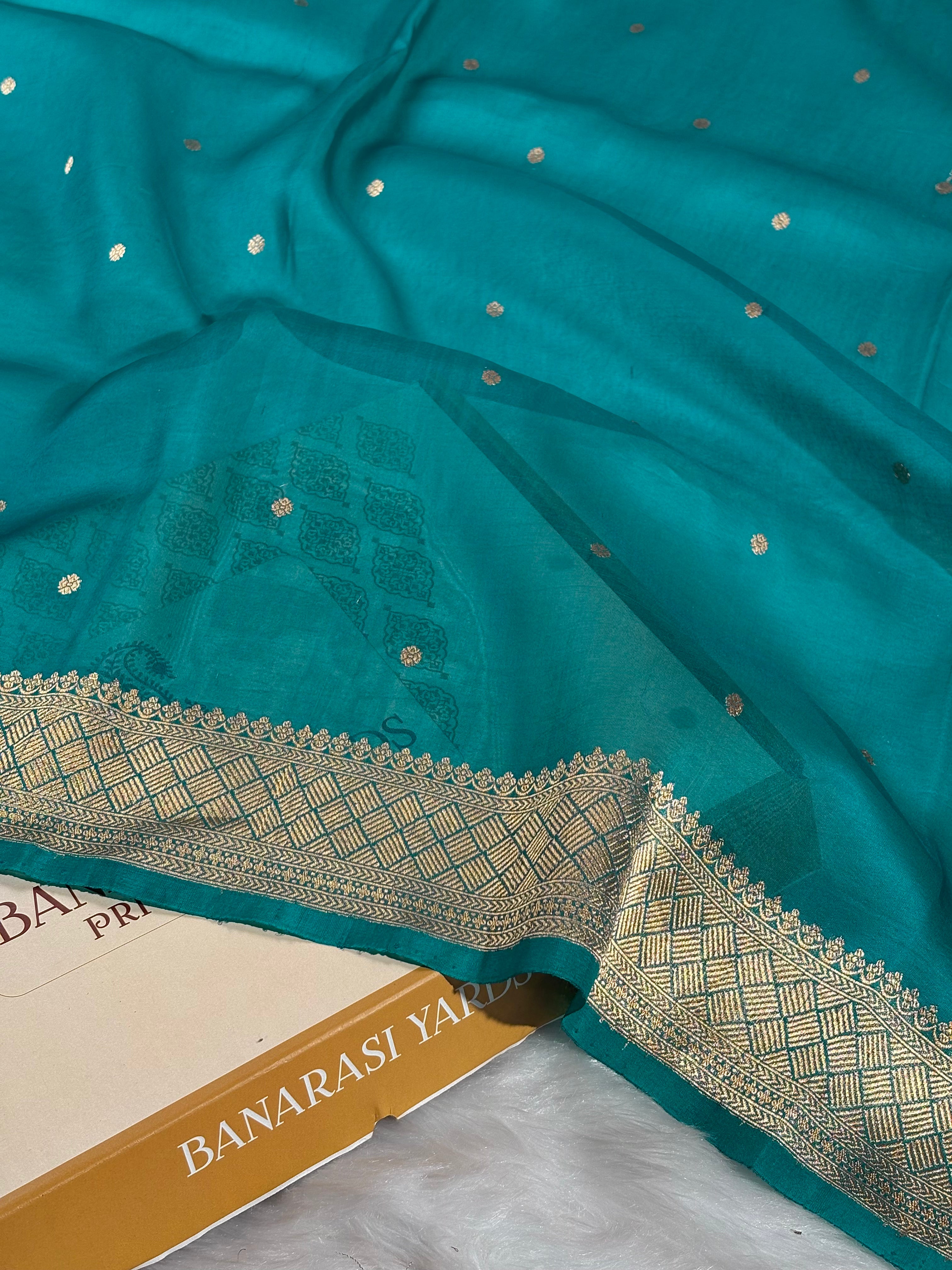 A teal green Banarasi silk saree with gold zari woven borders and small gold motifs, draped over a box with a white faux fur surface underneath.