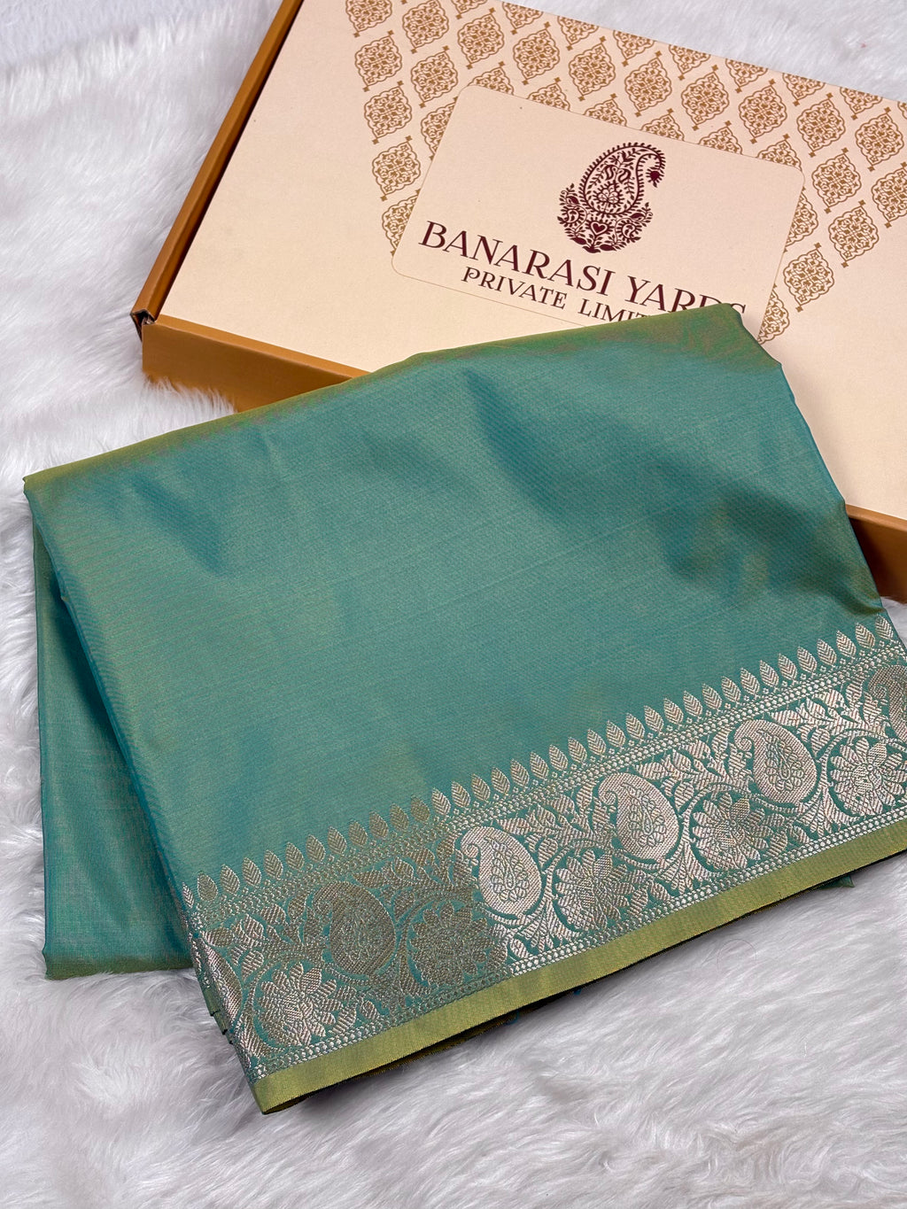 A folded sage green silk saree with a silver zari border, displayed next to its Banarasi Yards Private Limited box on a white faux fur background.