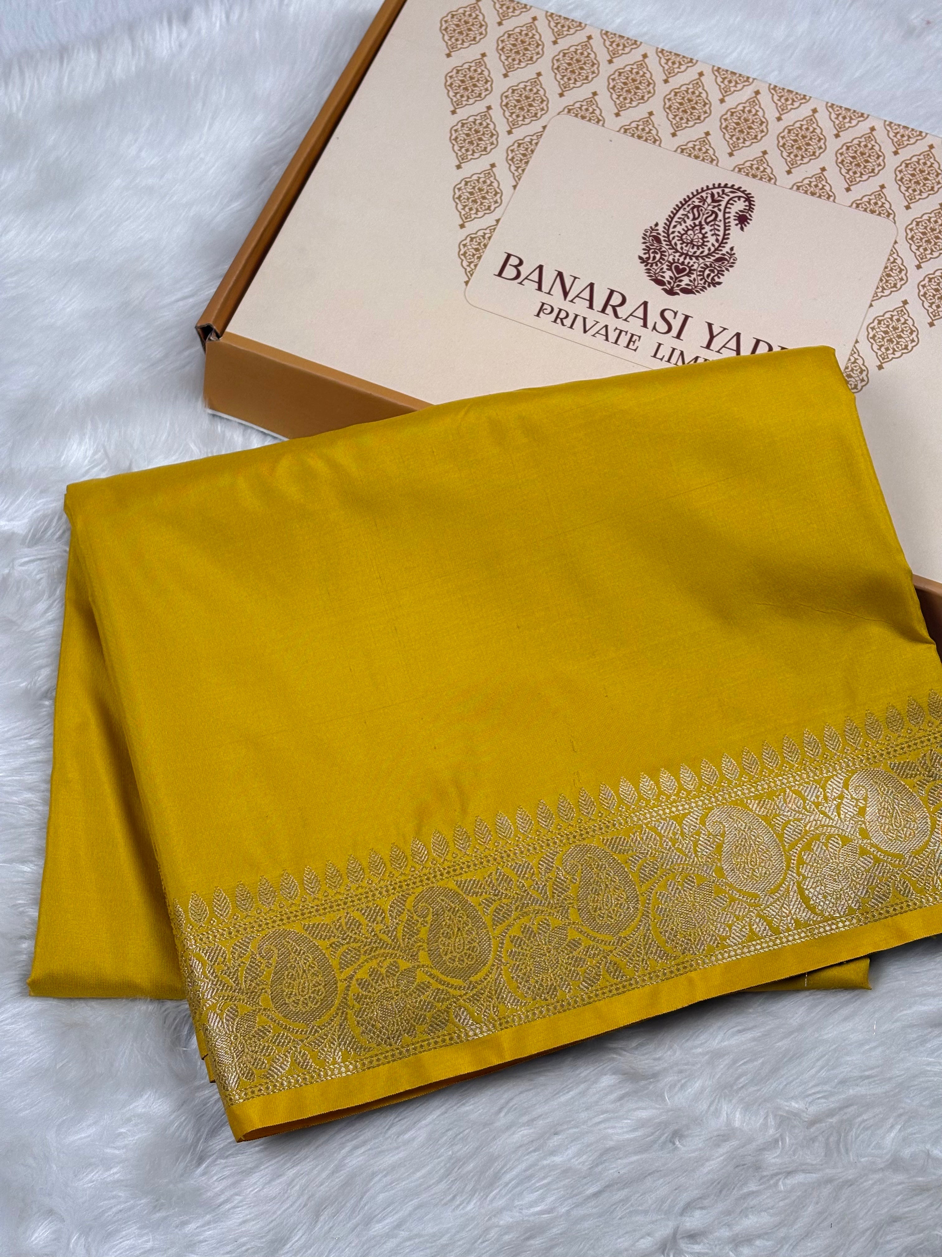 A folded mustard yellow Banarasi silk saree with a detailed gold zari border, displayed next to its branded packaging box on a white faux fur surface.