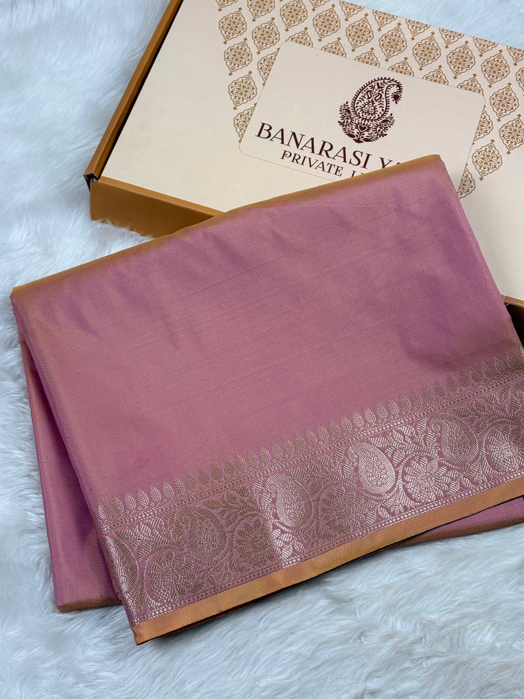 A folded dusty pink silk saree with a metallic gold border, displayed next to its branded 'Banarasi Yards Private Limited' gift box on a white faux fur surface.