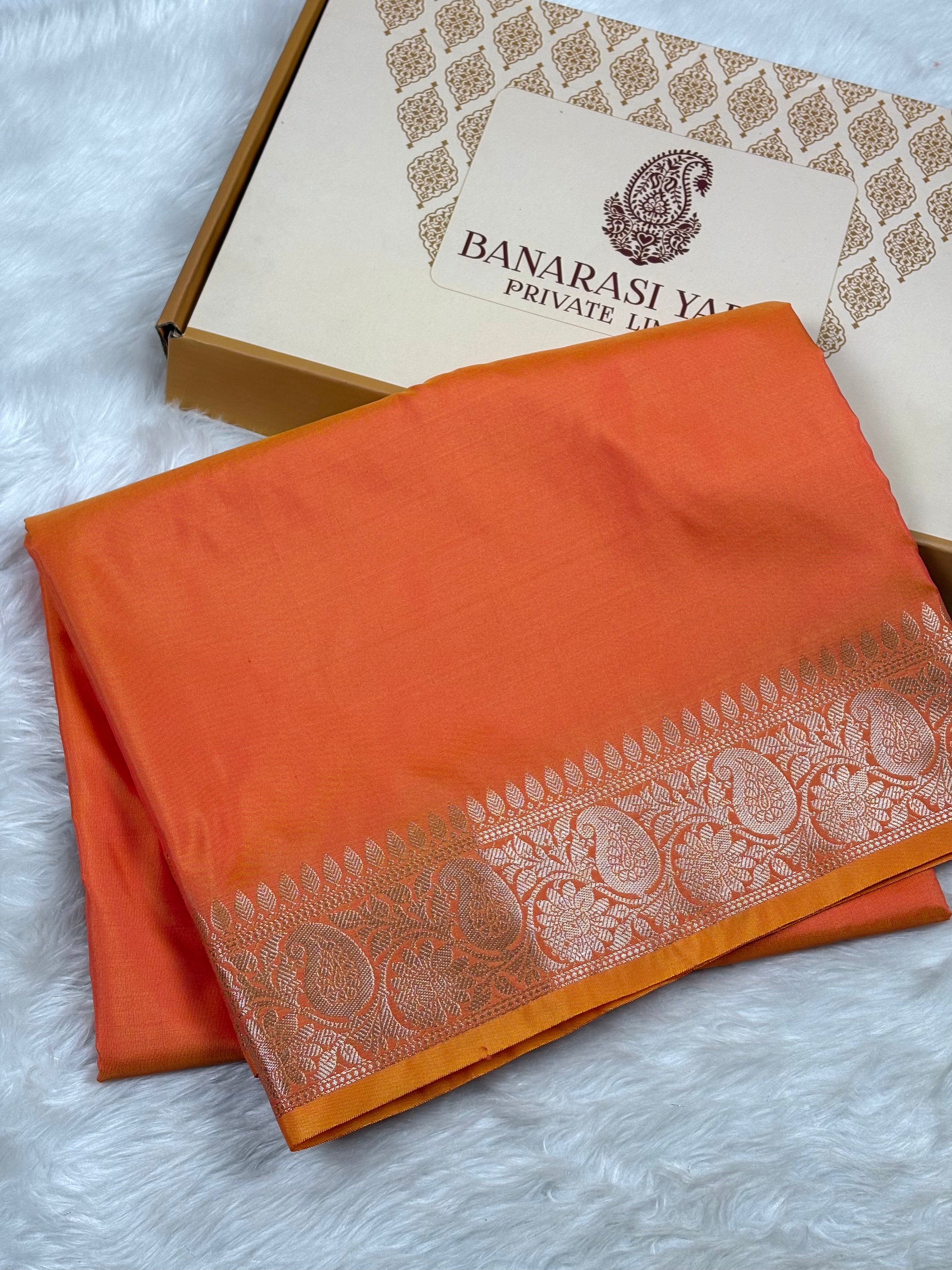 An orange Banarasi silk saree with a silver zari border, folded and displayed next to its branded packaging box on a white faux fur surface.