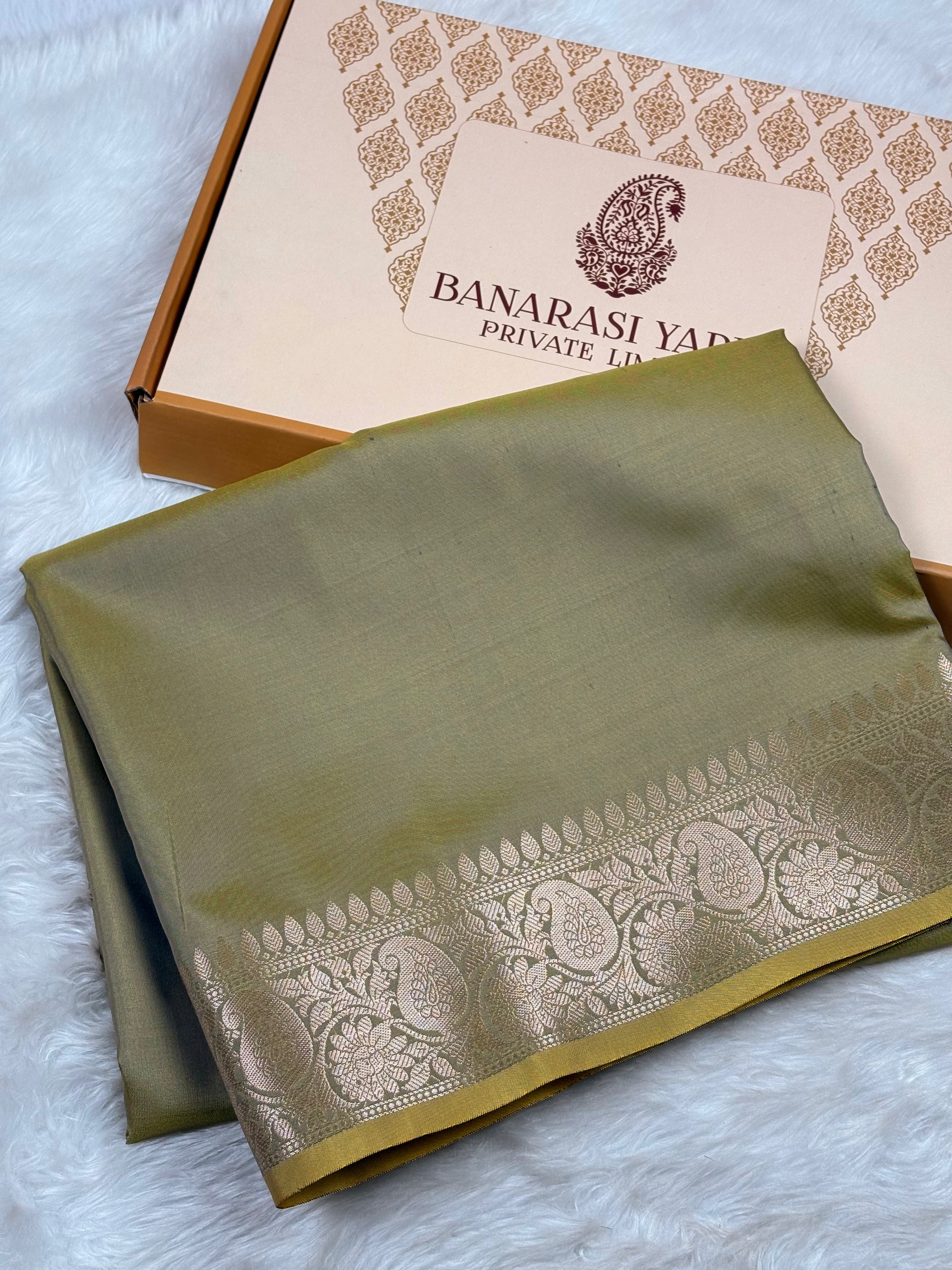 An olive green Banarasi silk saree with a gold zari border, folded and displayed next to its branded packaging box on a white faux fur surface.