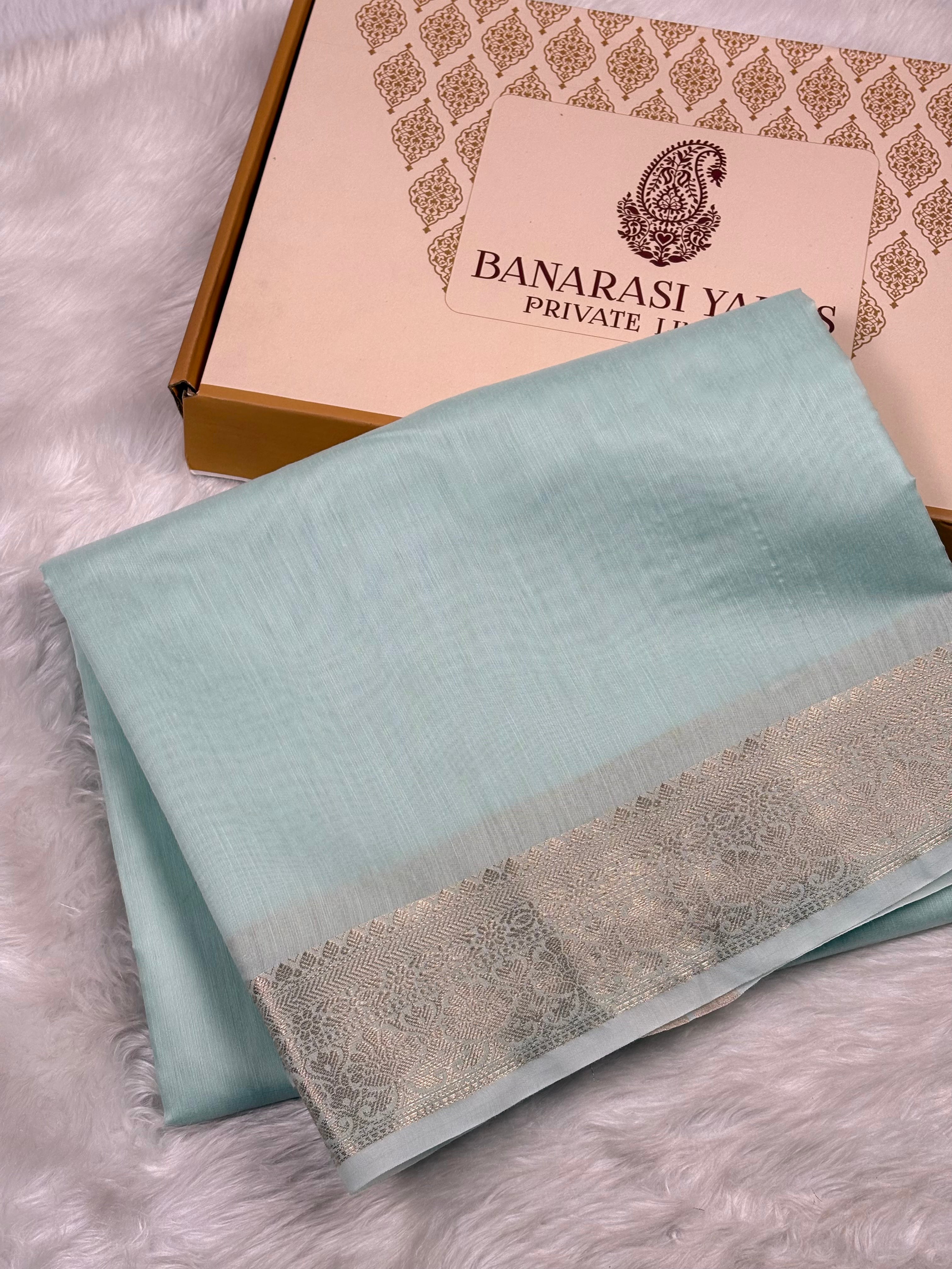 A folded light blue-green saree with a silver patterned border, displayed next to its Banarasi Yards Private Limited box on a white faux fur surface.