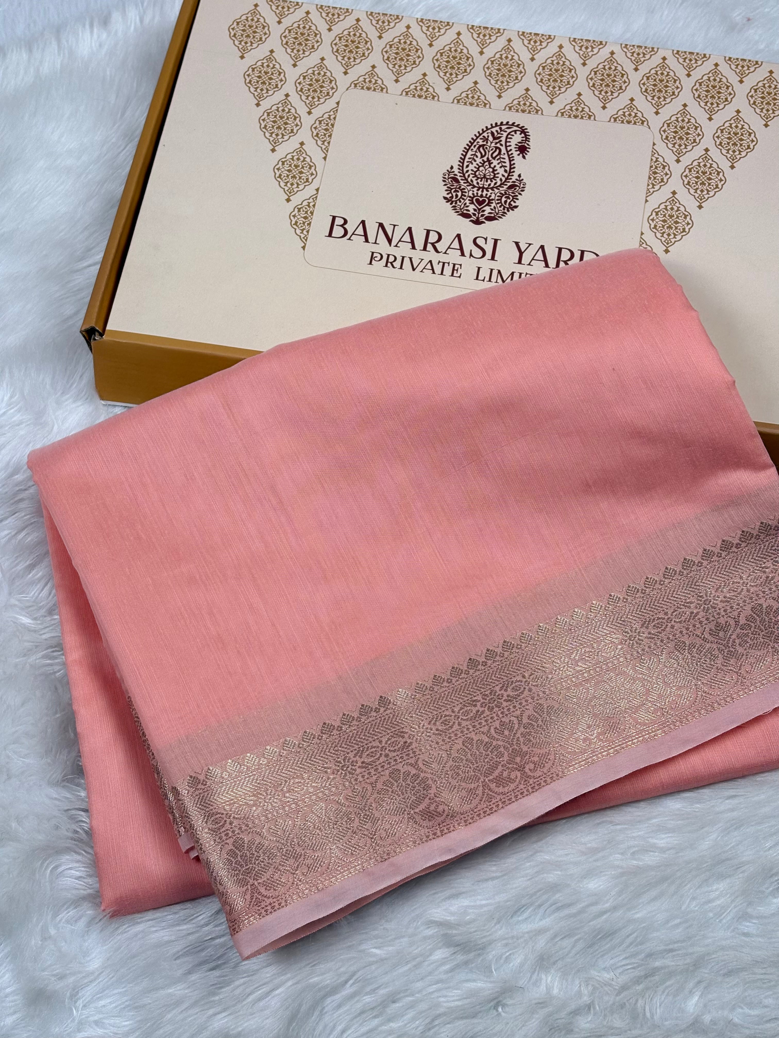 A folded dusty pink silk saree with a gold border, displayed next to its branded packaging box on a white faux fur surface.