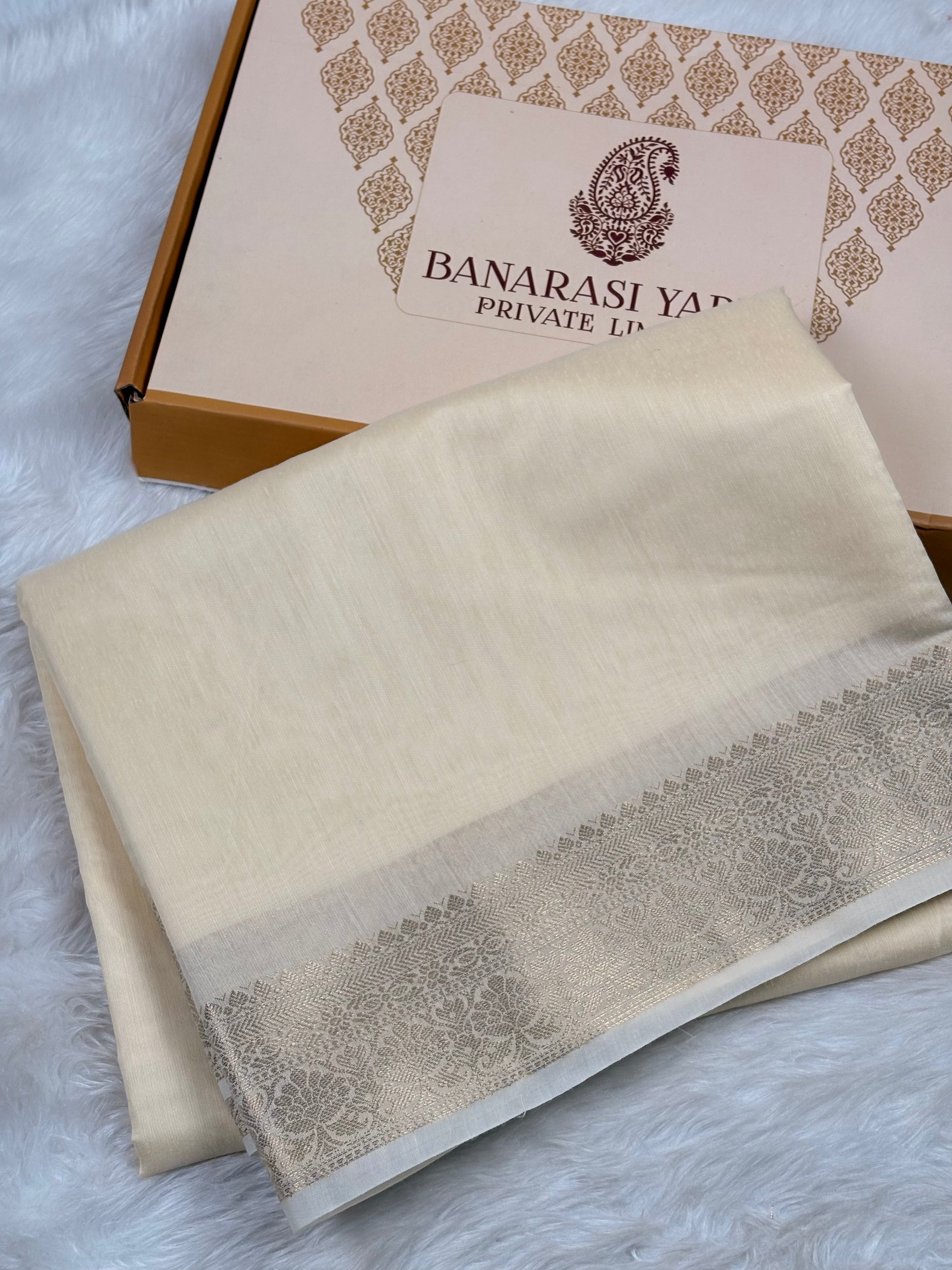 A folded cream-colored Banarasi silk saree with a gold border, displayed next to its branded packaging box on a white faux fur surface.