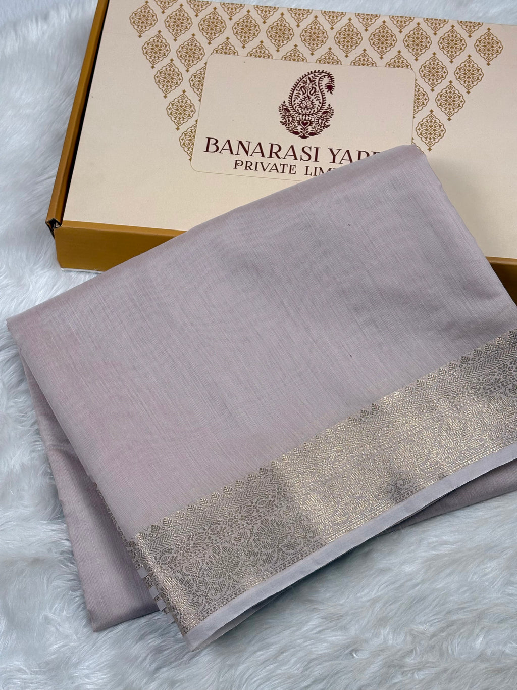 A folded light lavender-gray saree with a gold border, displayed next to its branded packaging box labeled 'BANARASI YARDS PRIVATE LIMITED'.