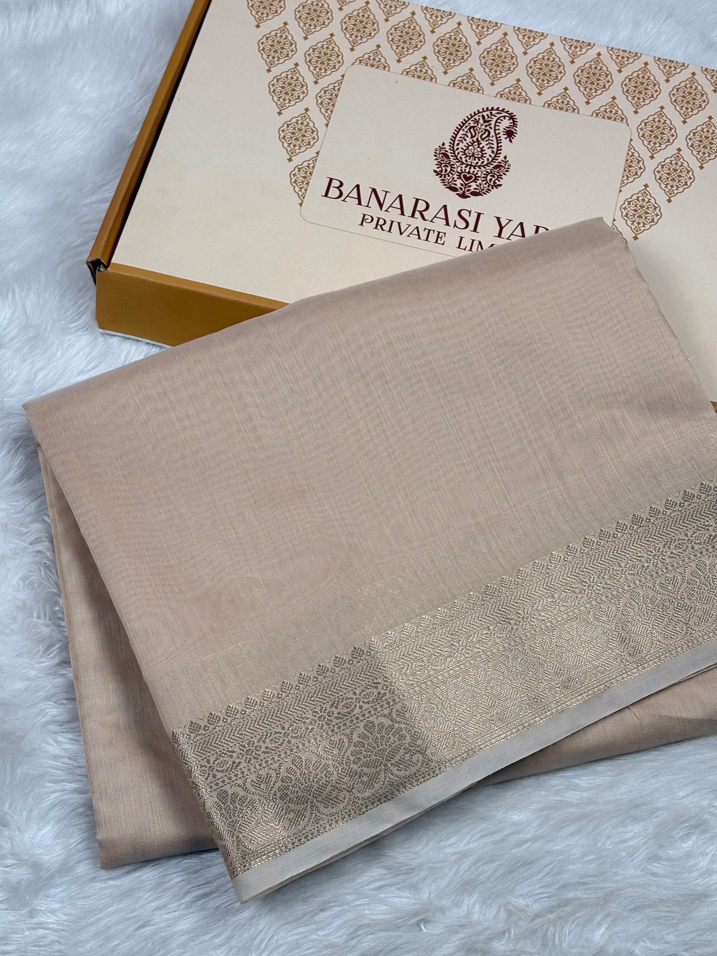 A folded beige Banarasi silk saree with a gold border, displayed next to its branded packaging box on a white faux fur surface.