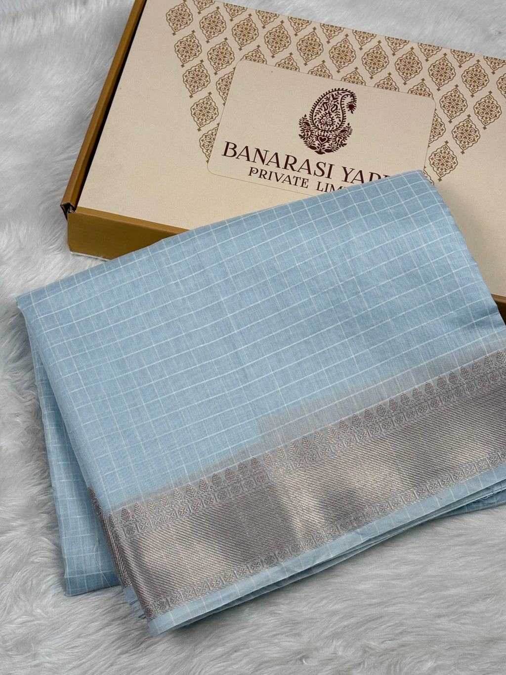 A folded light blue Banarasi silk saree with a silver border, displayed next to its branded packaging box on a white faux fur surface.