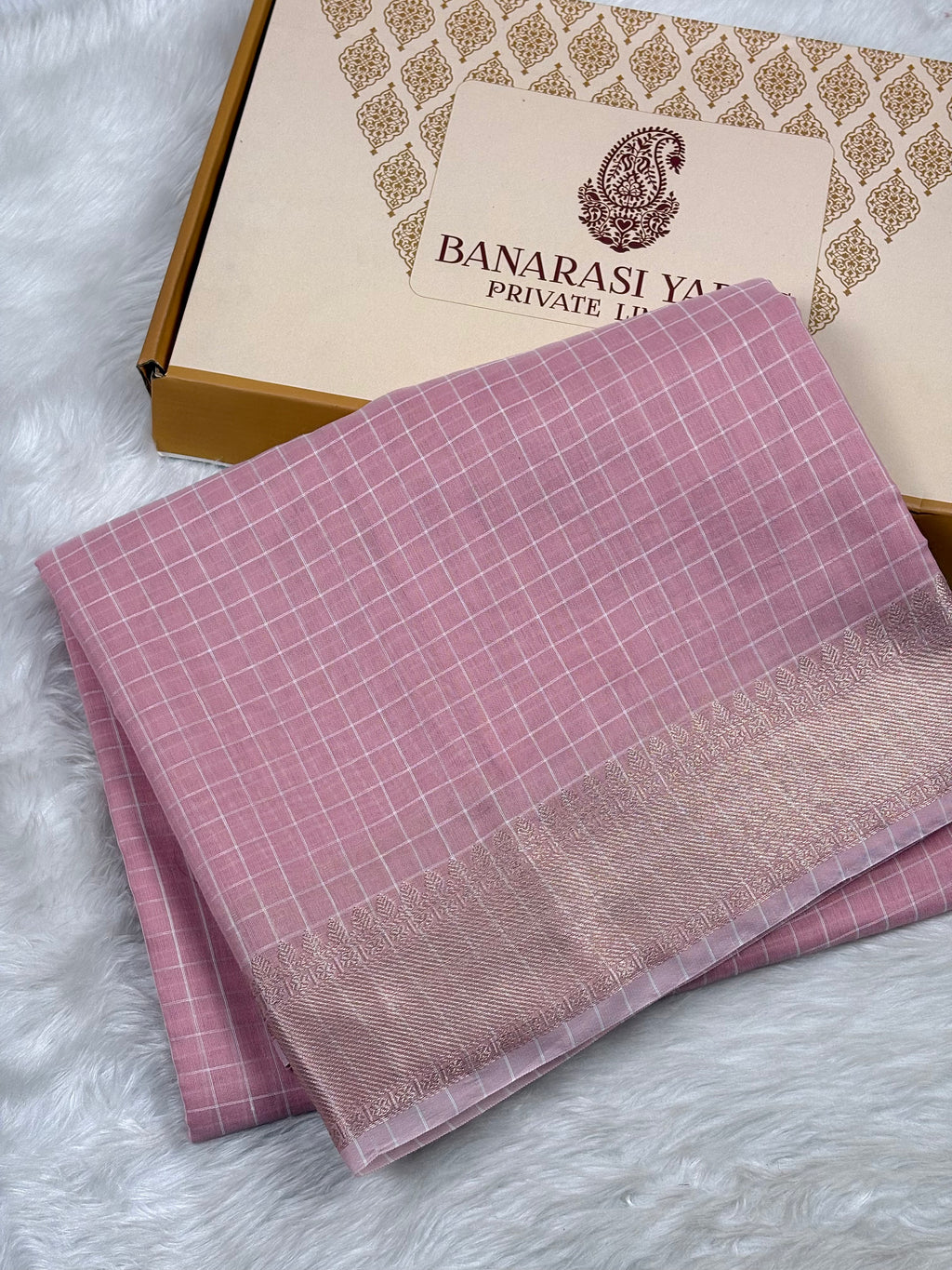 A folded pink Banarasi silk saree with a subtle grid pattern and a zari border, displayed next to its branded packaging box on a white faux fur surface.