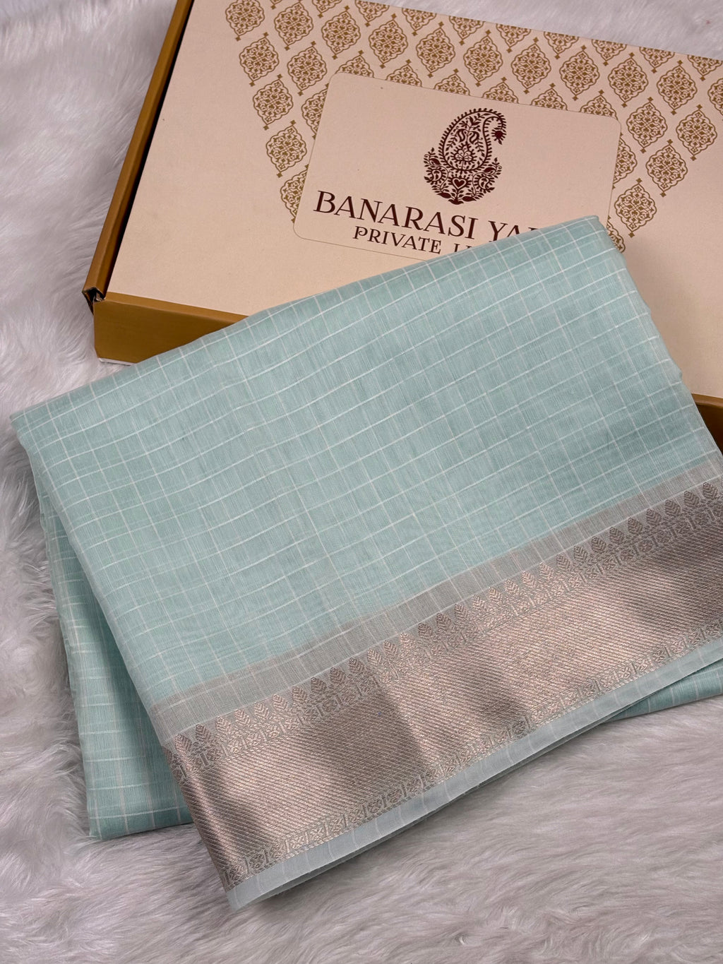 A folded light blue saree with a silver border, displayed next to its Banarasi Yards Private Limited gift box on a white faux fur surface.