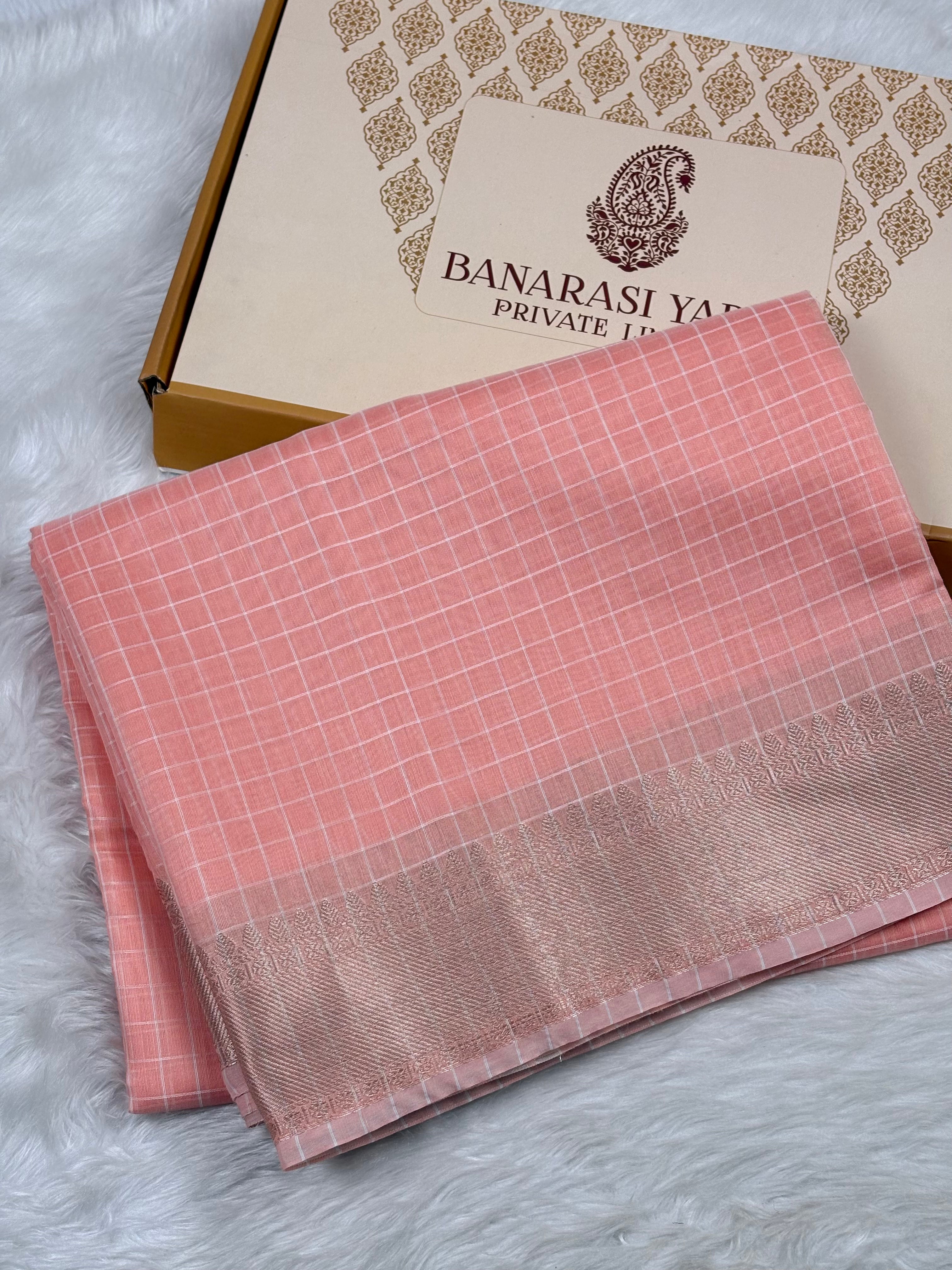 A folded pink Banarasi silk saree with a subtle grid pattern and a gold border, displayed next to its branded packaging box on a white faux fur surface.