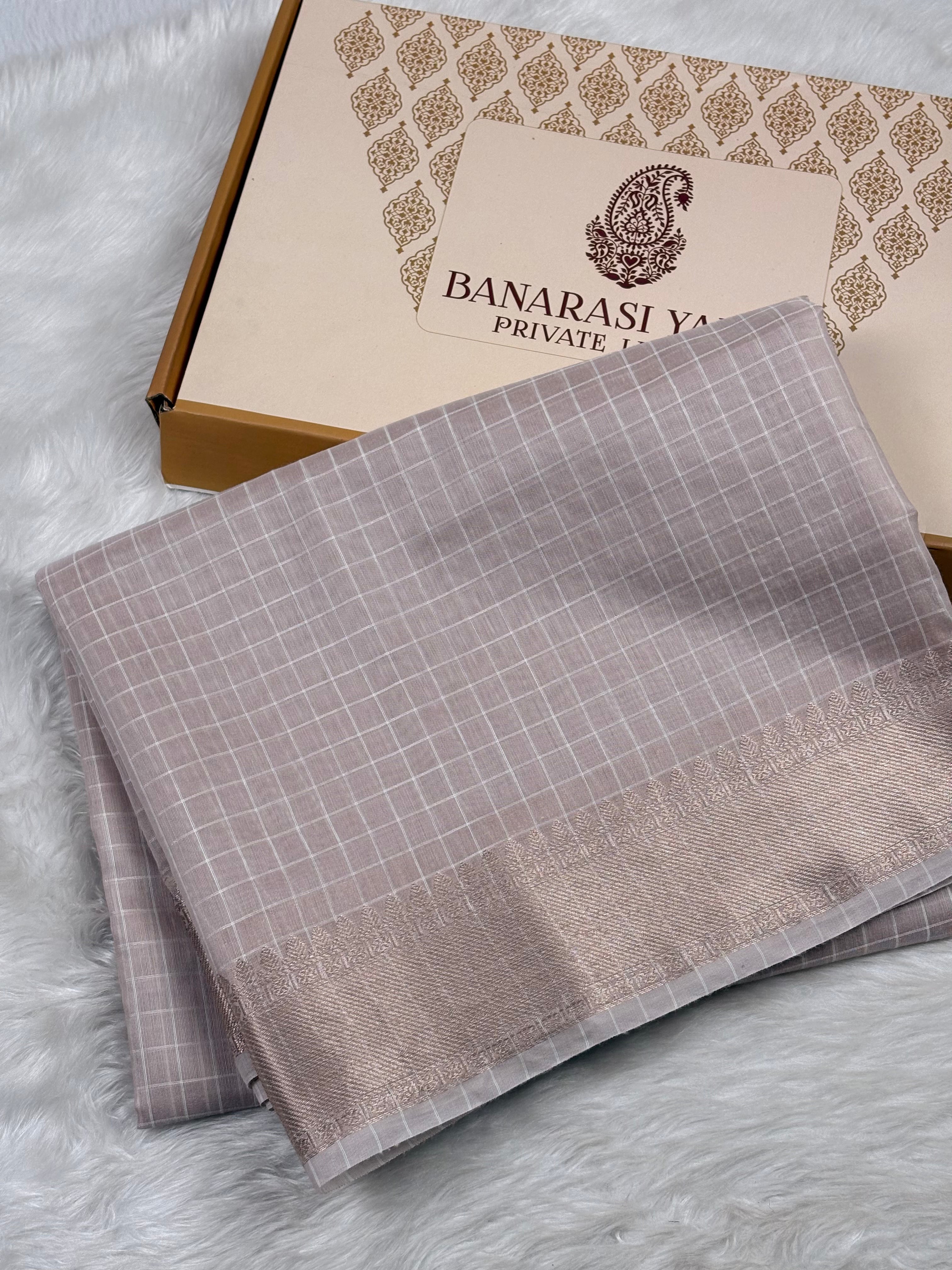 A folded Banarasi silk saree in a muted taupe-gray color with a fine white grid pattern and a wide gold border, displayed next to its branded packaging box on a white faux fur surface.