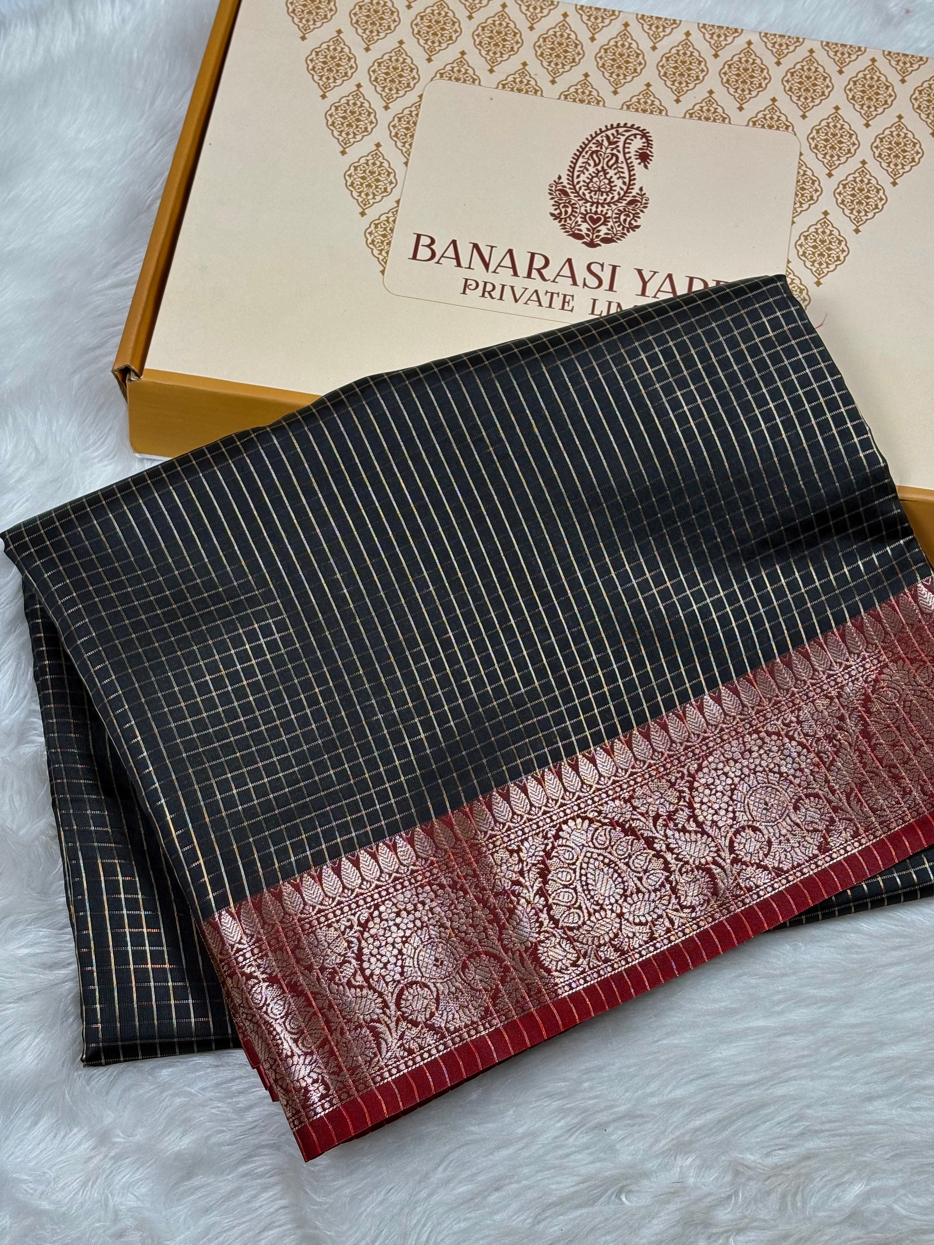 A folded black Banarasi silk saree with gold zari stripes and a red border with intricate gold zari work, displayed next to its branded gift box on a white faux fur surface.