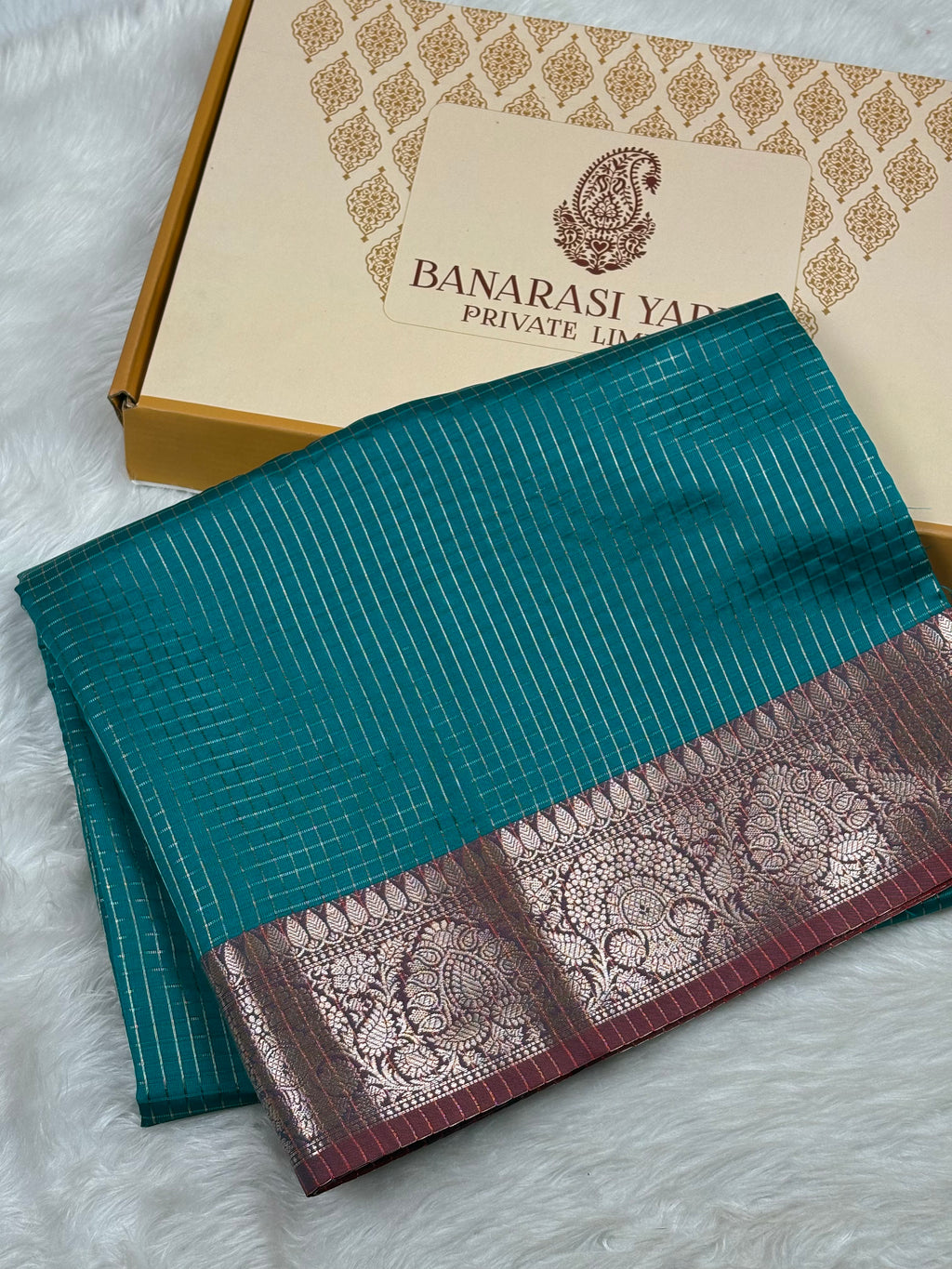 A folded teal blue saree with a gold border and a deep pinkish-purple border, displayed next to its branded packaging box on a white faux fur surface.