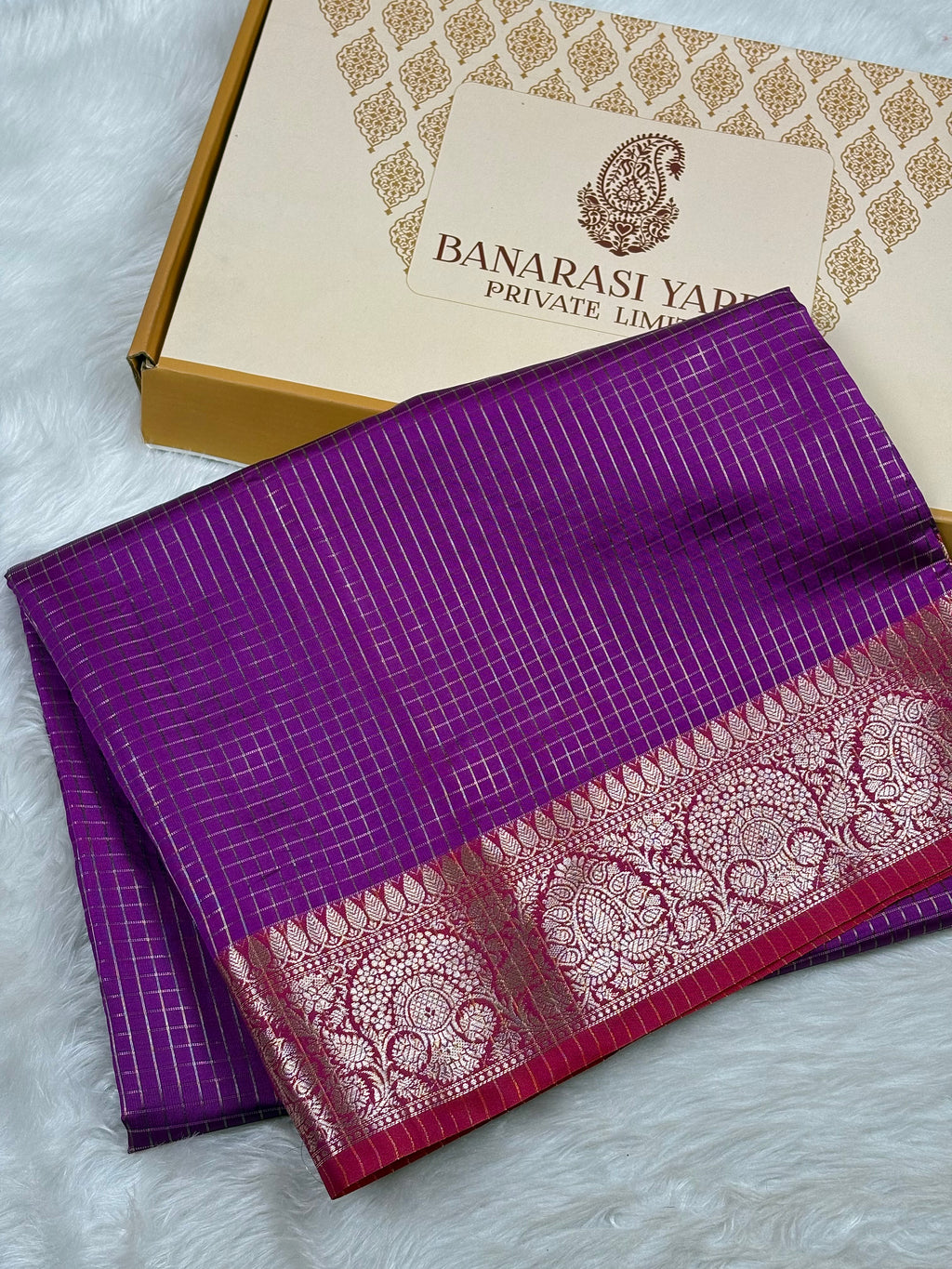 A folded purple Banarasi silk saree with a detailed gold zari border, displayed next to its branded packaging box on a white faux fur surface.