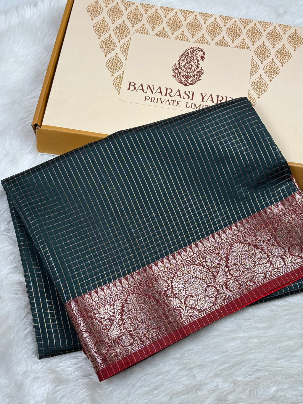 A folded dark teal-green saree with fine white stripes and a wide red border featuring intricate gold zari weaving, displayed next to its branded packaging box on a white faux fur surface.
